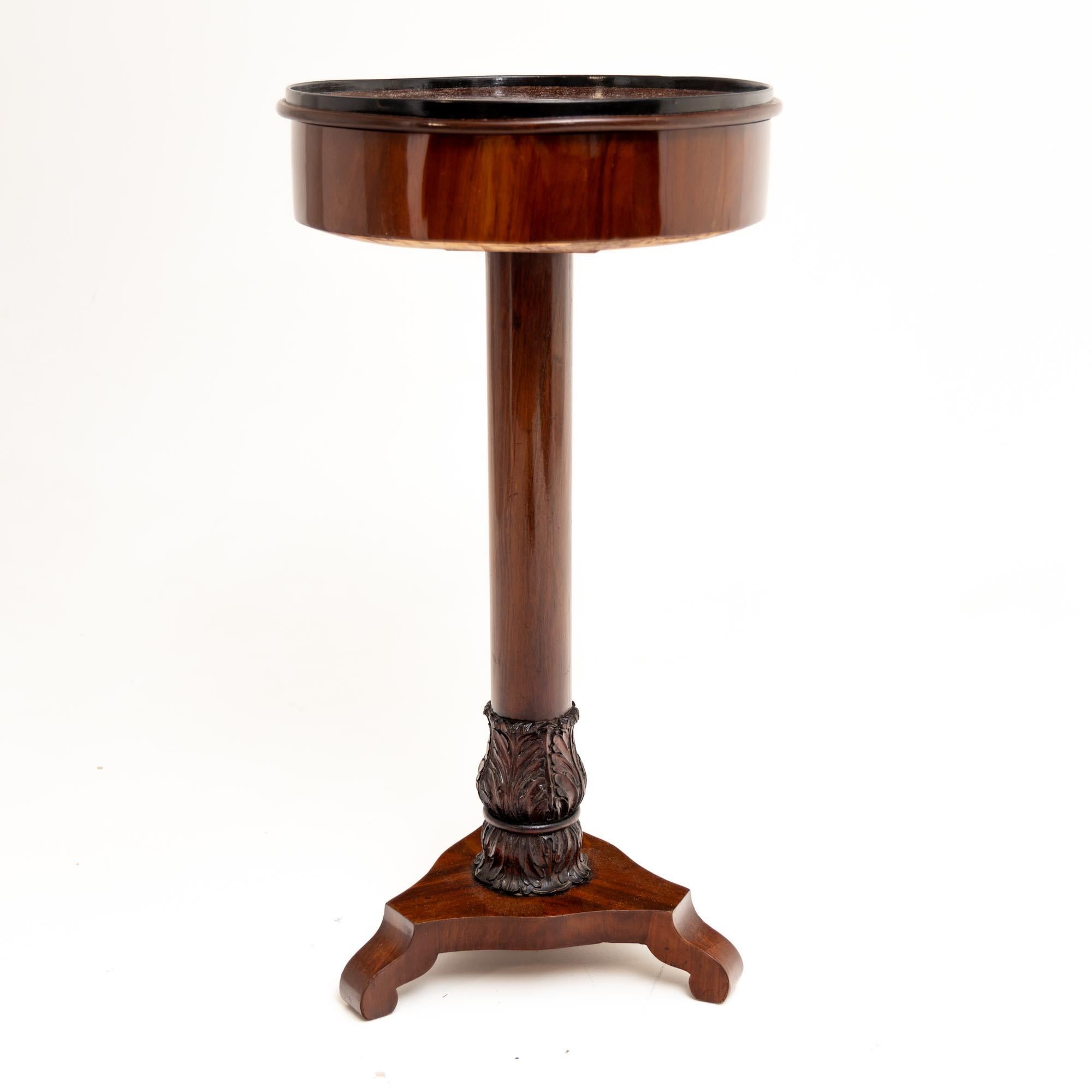 Small side table on a trefoil base with volute-shaped legs and carved leaf decorations in the lower section. Above it rises the smooth central column and the round table top with smooth frame. The lid with ebonised edge is removable and reveals the