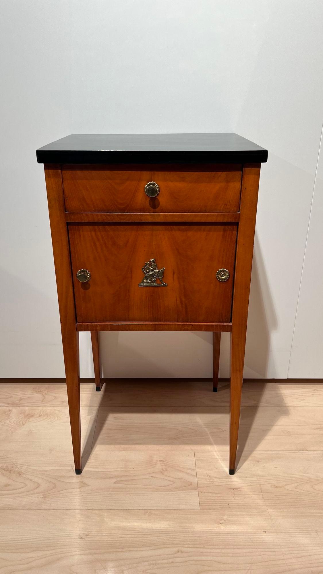 Fine Biedermeier small furniture or pillar cabinet from southern Germany around 1820.

Solid cherry wood legs and veneered body. Ebonized top plate.
One drawer at the top. Below one door with a beautiful central brass ornament of a woman playing the