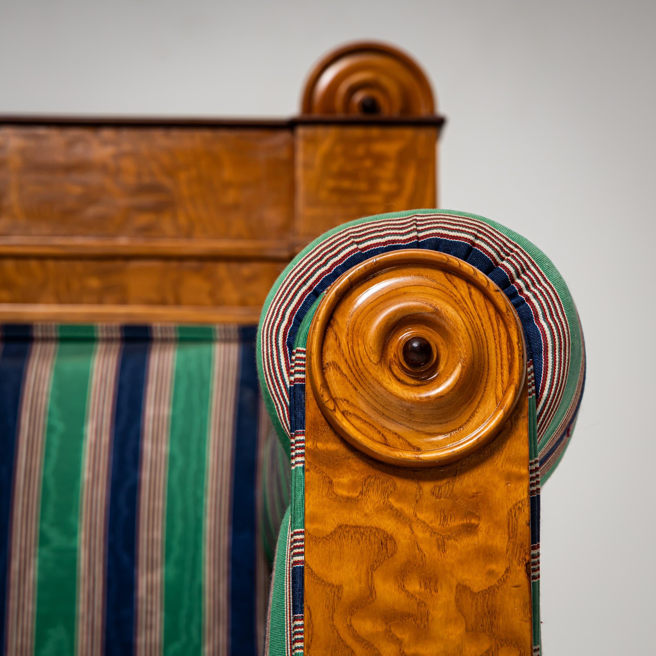 Elegante sofá Biedermeier con armazón chapado en madera de fresno, fabricado hacia 1830. El asiento, los reposabrazos, el respaldo y la parte trasera están totalmente tapizados en una tela de rayas verdes y azules. Su respaldo recto y su base