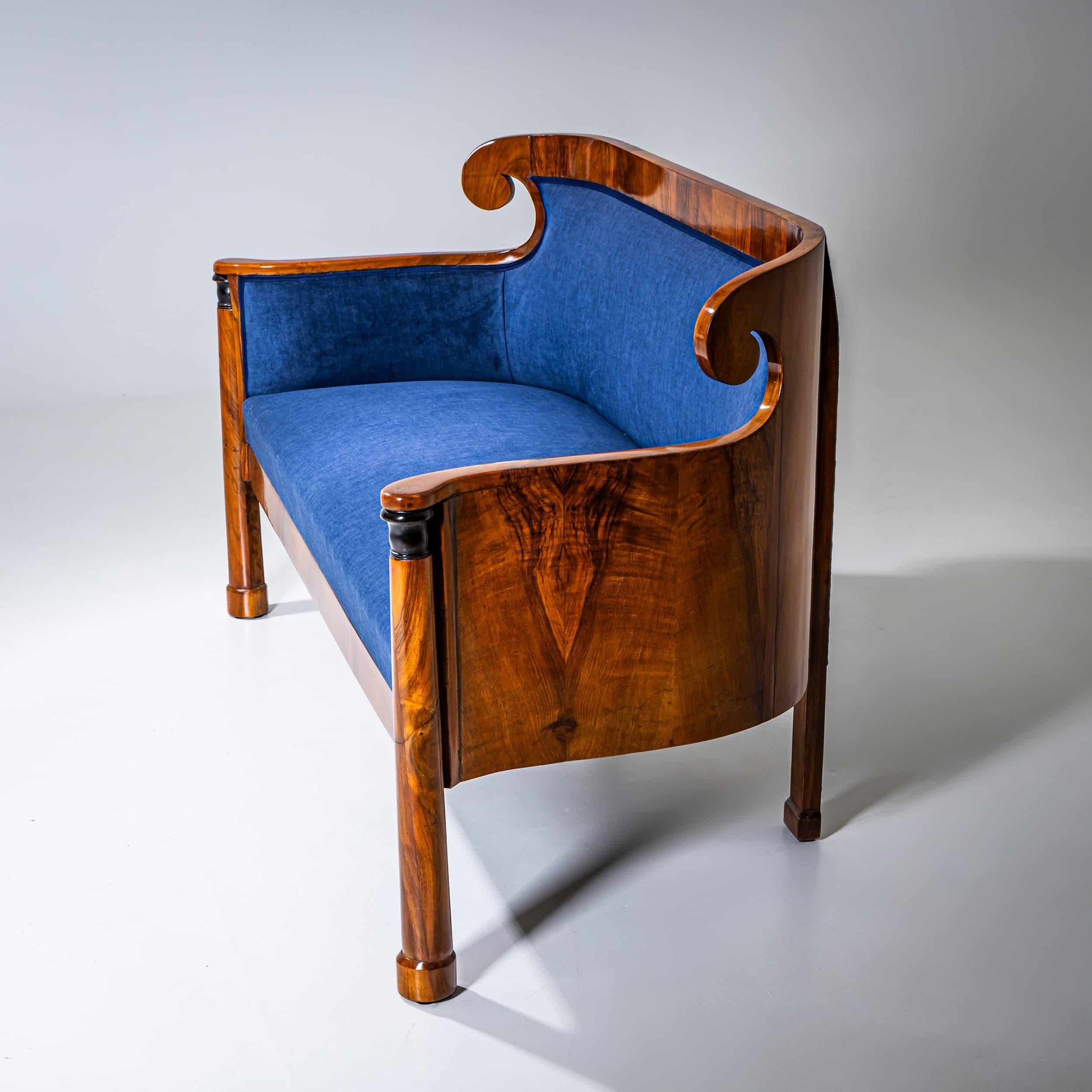 Divano a due posti Biedermeier impiallacciato in noce, poggiante su gambe a colonna con capitelli ebanizzati. Lo schienale dolcemente curvo termina con volute stilizzate. Il divano è stato rivestito di recente in tessuto blu scuro e lucidato a mano.