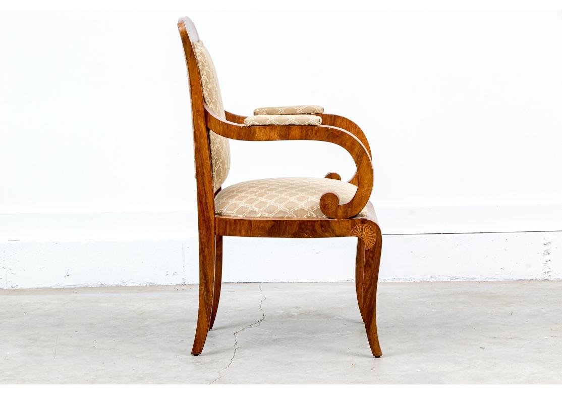 Sillón tapizado de estilo Biedermeier con incrustaciones de marquetería en la crestería y el delantal. La silla está tapizada en un tejido neutro con motivos geométricos y doble ribete. Los reposabrazos, con atrevidas volutas, están cubiertos de