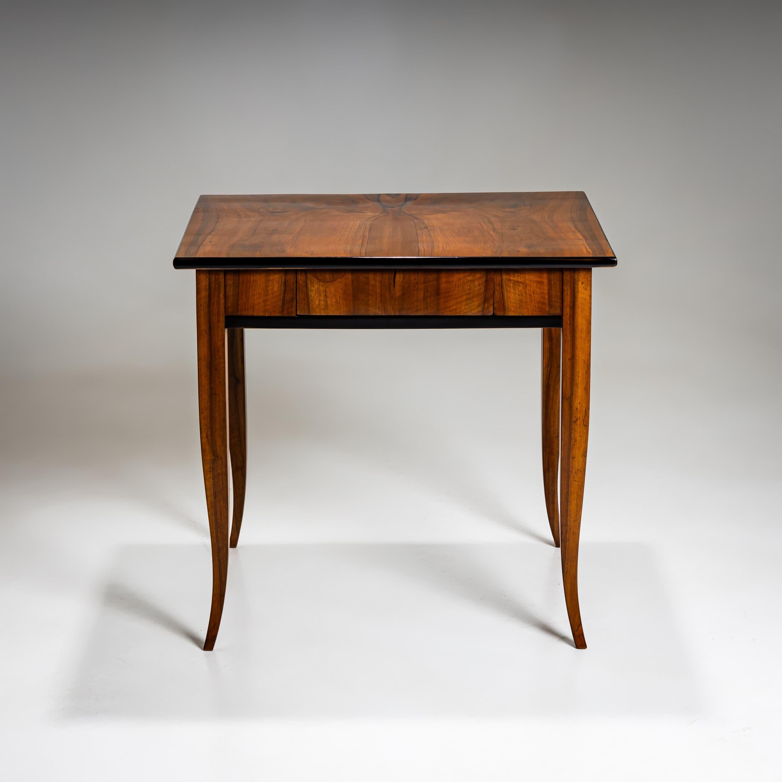 Elegant Biedermeier table featuring four gracefully curved legs and a small drawer. The surface is walnut-veneered, with ebonized edges that create a refined contrast. The table has been professionally restored and hand-polished.