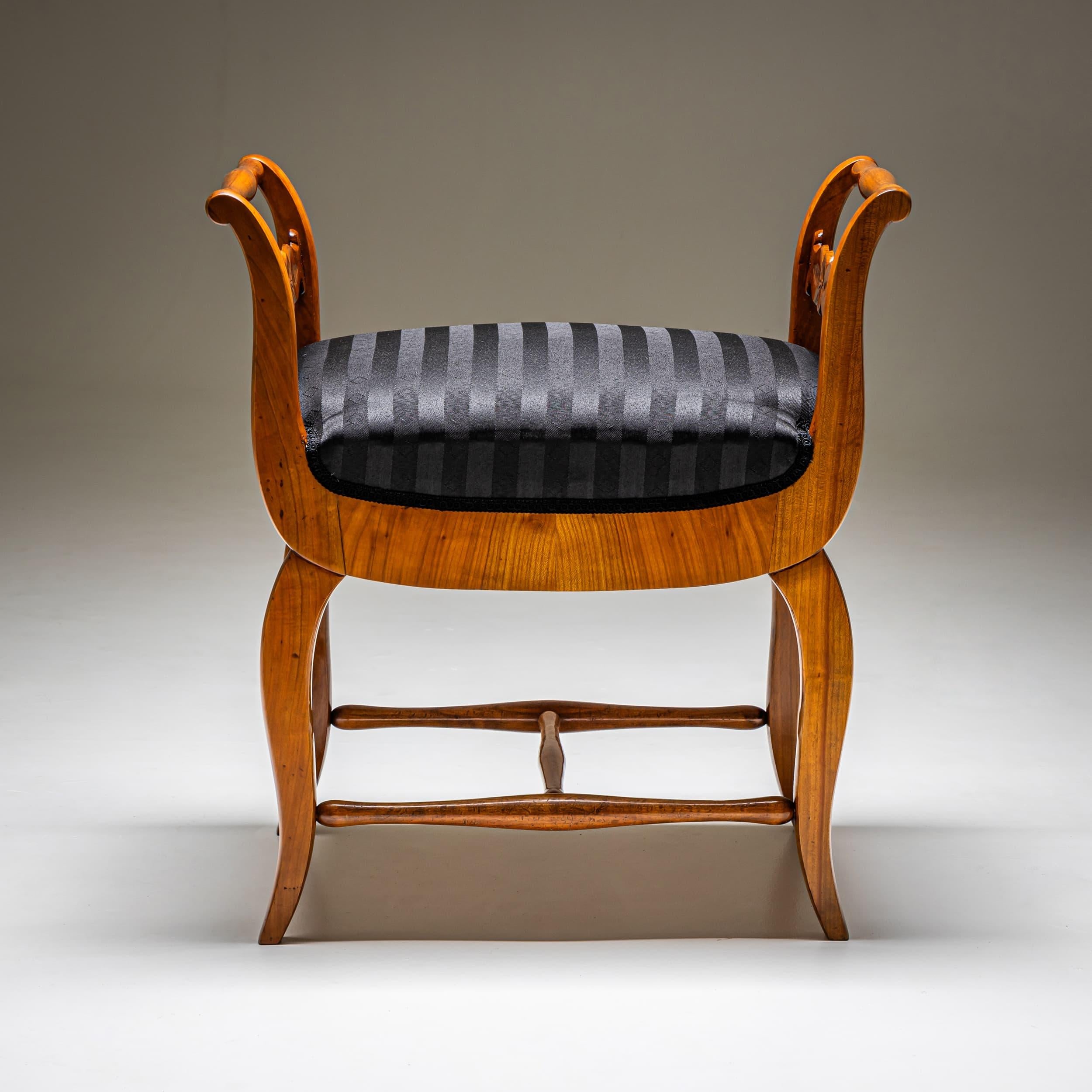 Biedermeier Tabouret aus Kirschholz mit H-förmiger Bahre, die die geschwungenen Beine verbindet. Die Armlehnen sind durch einen Querbalken verbunden, der mit stilisierten Blattmotiven verziert ist. Der Sitz wurde neu gepolstert und mit einem schwarz