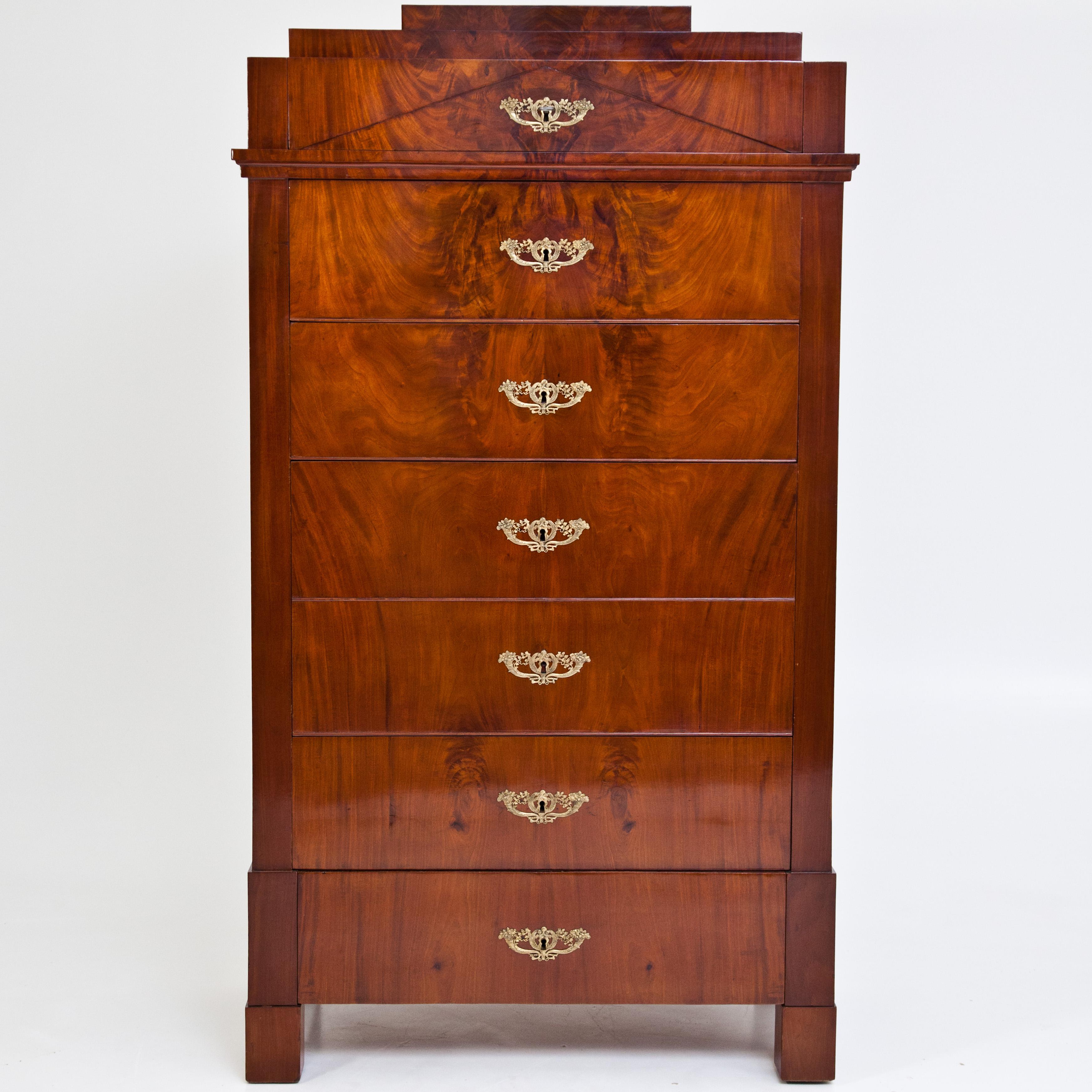Tall chest of drawers on square feet with five drawers and a large optically blended drawer in the centre. The stepped top with triangular filling and profiled cornice strip below the first drawer. Solid mahogany and veneered. Very good, hand