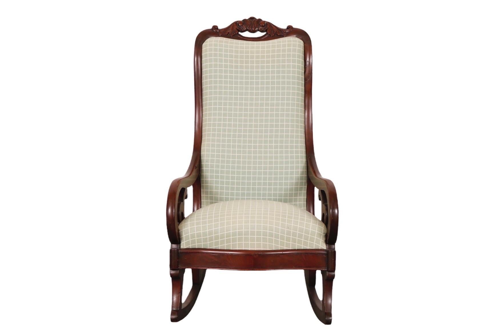 A Biedermeier rocking chair made of mahogany. Carved with a pierced, scrolled acanthus crest rail and scrolled arms, the seat back has a serpentine curved edge mirrored in the bow front skirt. Upholstered in a sage green fabric with a white grid