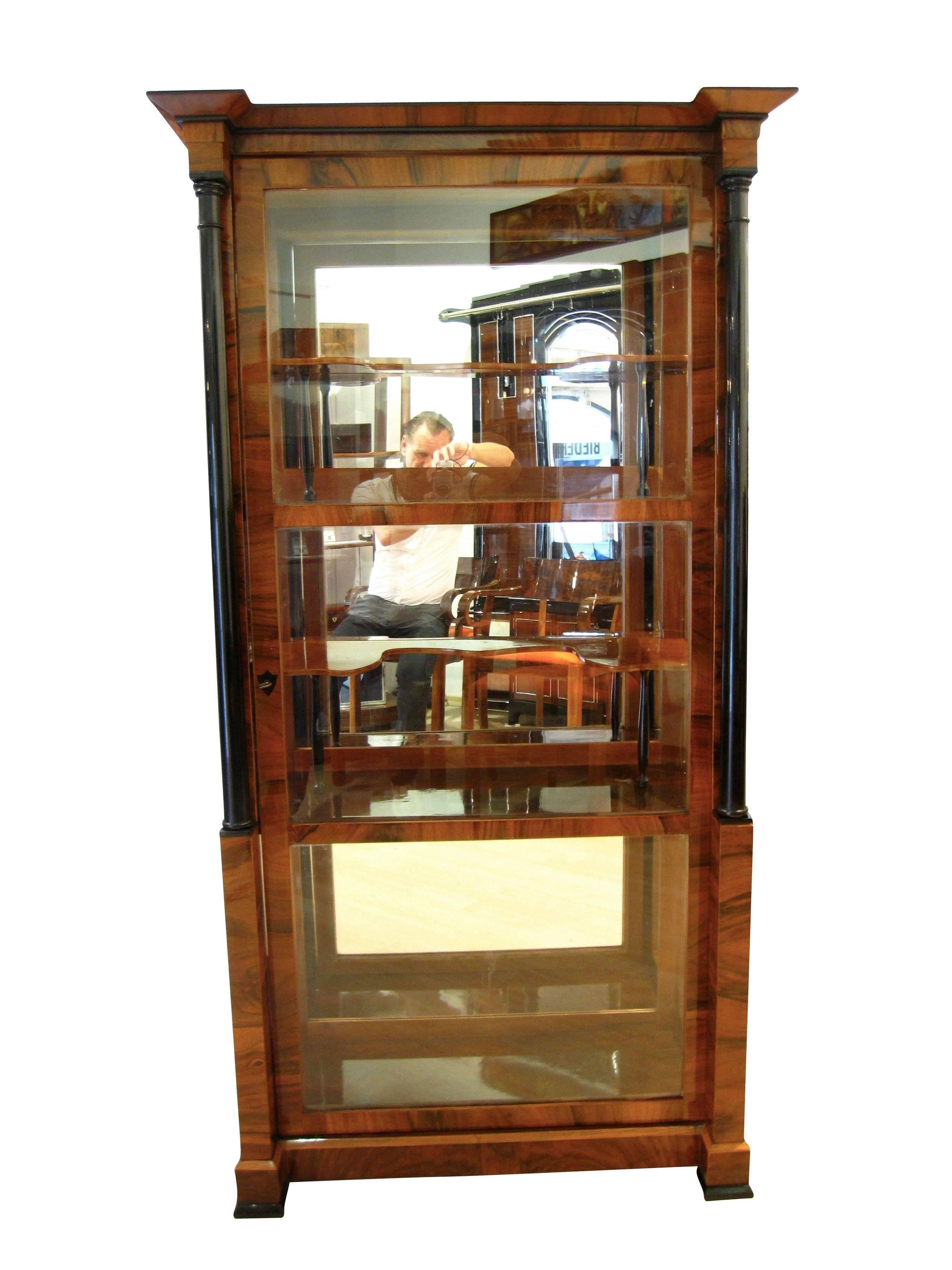 Beautiful, classic Vitrine with glass windows on three sides form the 19th century. The wood is walnut veneer and solid wood and it has two ebonized full columns. 
Inside there are two shelves and in between two smaller curved middle shelves with