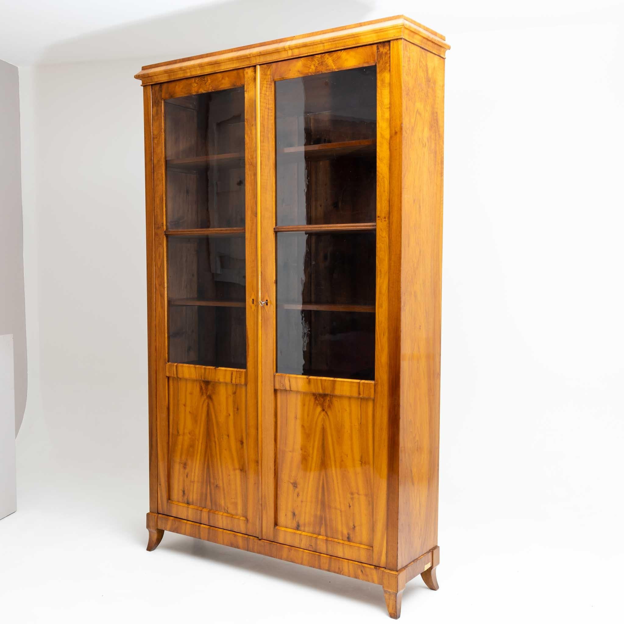 Two-door Biedermeier bookcase on small flared sabre legs with profiled flat cornice. The doors are two-thirds glazed. The cabinet has a corpus depth of 33 cm and is veneered in walnut. The interior consists of five shelves. The cabinet has been