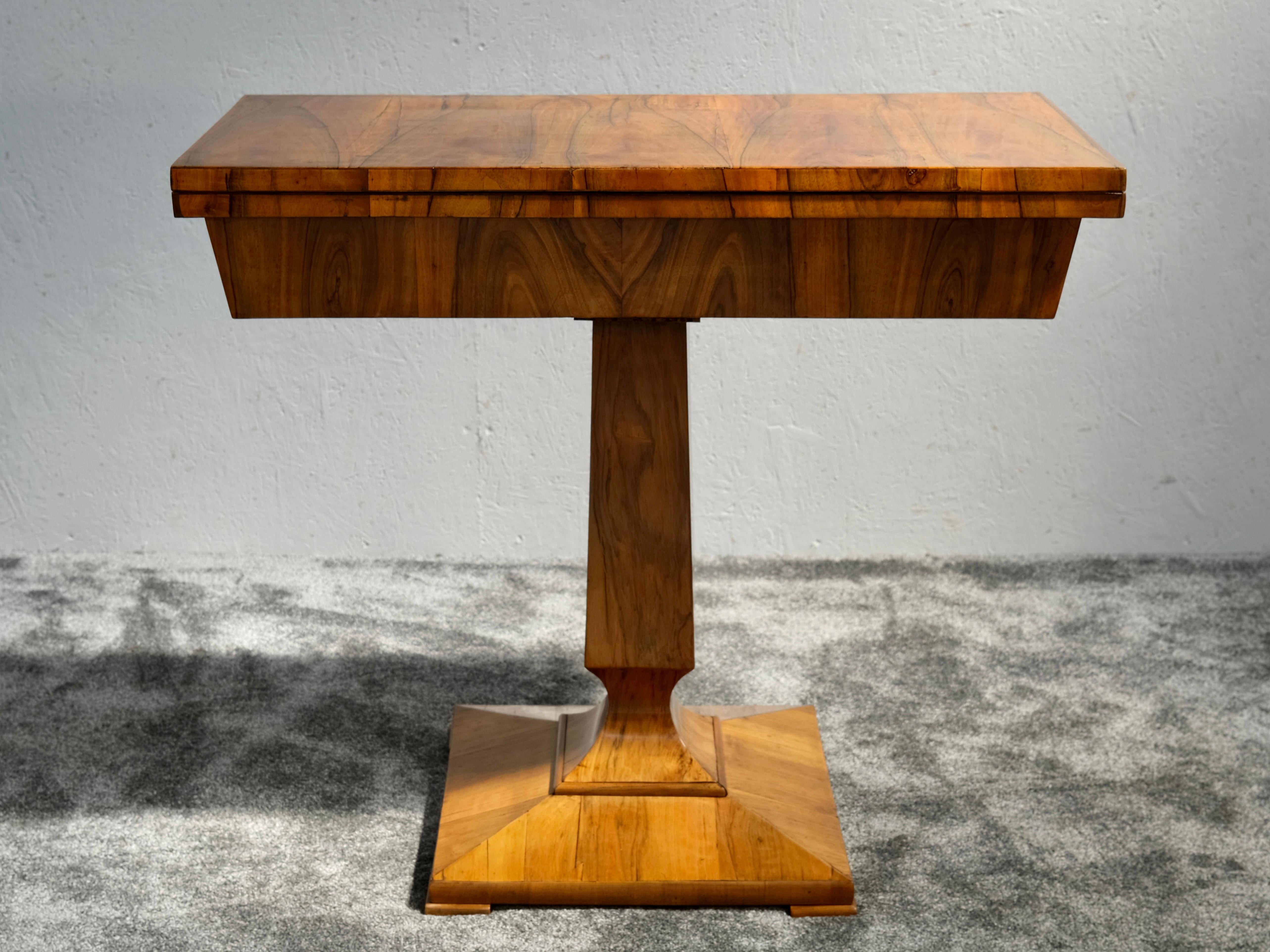 Esta mesa de juego abatible Biedermeier de hacia 1850 muestra la refinada elegancia del diseño centroeuropeo de principios del siglo XIX. Fabricada en madera blanda chapada en rico nogal, la pieza presenta una base de pedestal con un perfil cónico