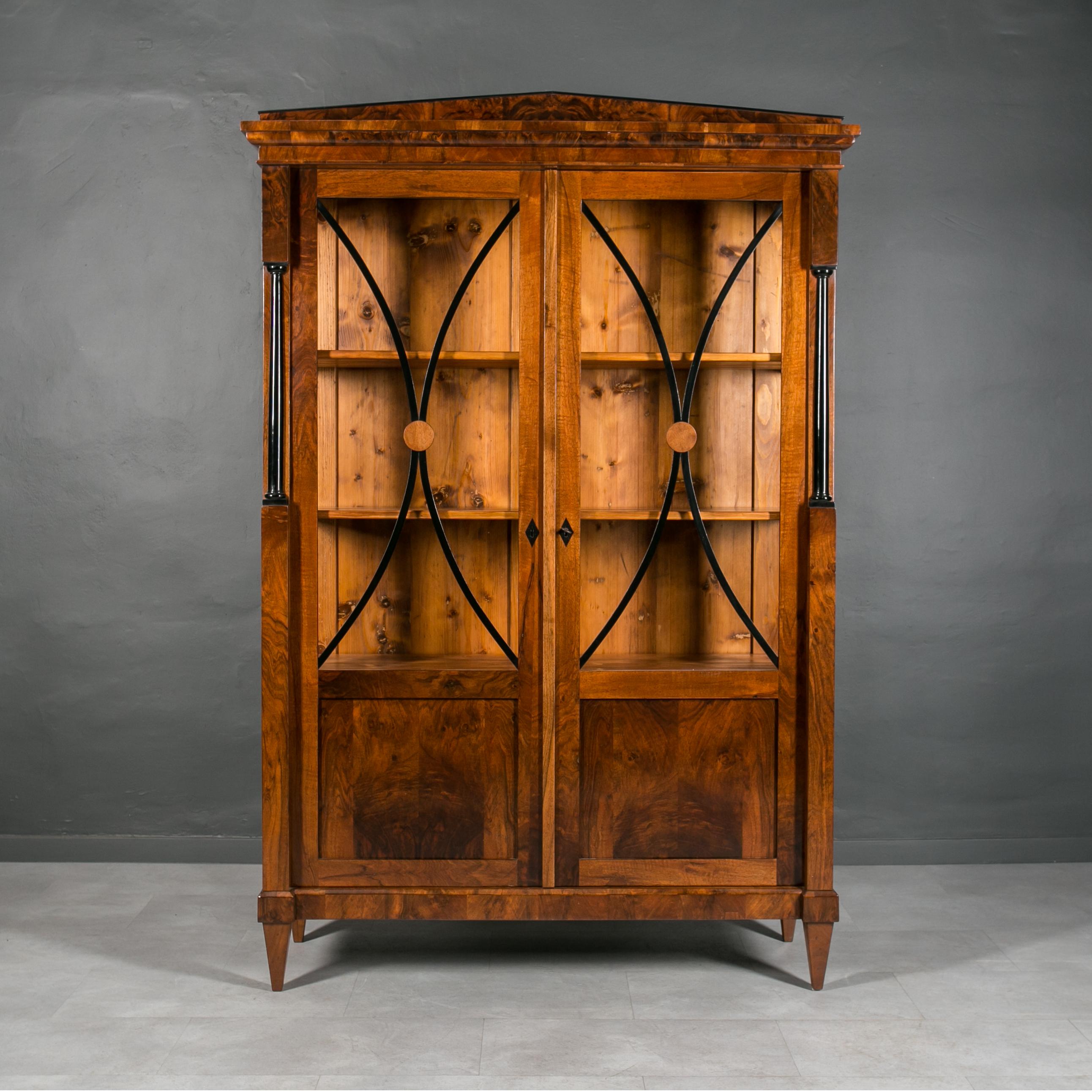 Diese schöne Vitrine stammt aus Deutschland und wurde im 19. Jahrhundert (Biedermeier) hergestellt. Die Konstruktion besteht aus Nadelholz, das mit schönem und reichem Nussbaumfurnier furniert ist. Hinter den mit einem Schlüssel abschließbaren Türen