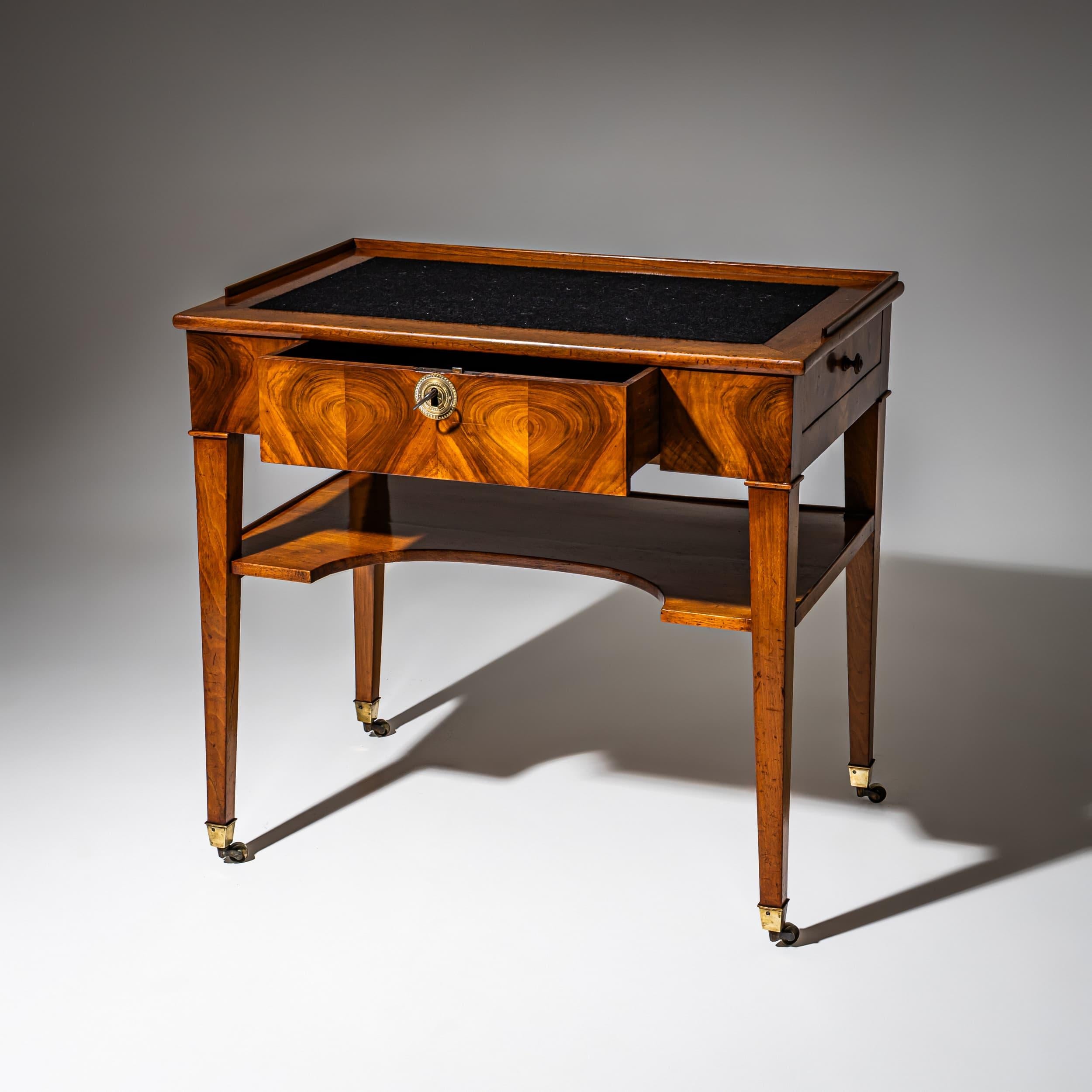 Small Biedermeier writing desk in walnut, raised on tapered square legs with brass sabots and casters. The desk features a central drawer with a large circular brass escutcheon and lock. The writing surface is covered in black felt and bordered on
