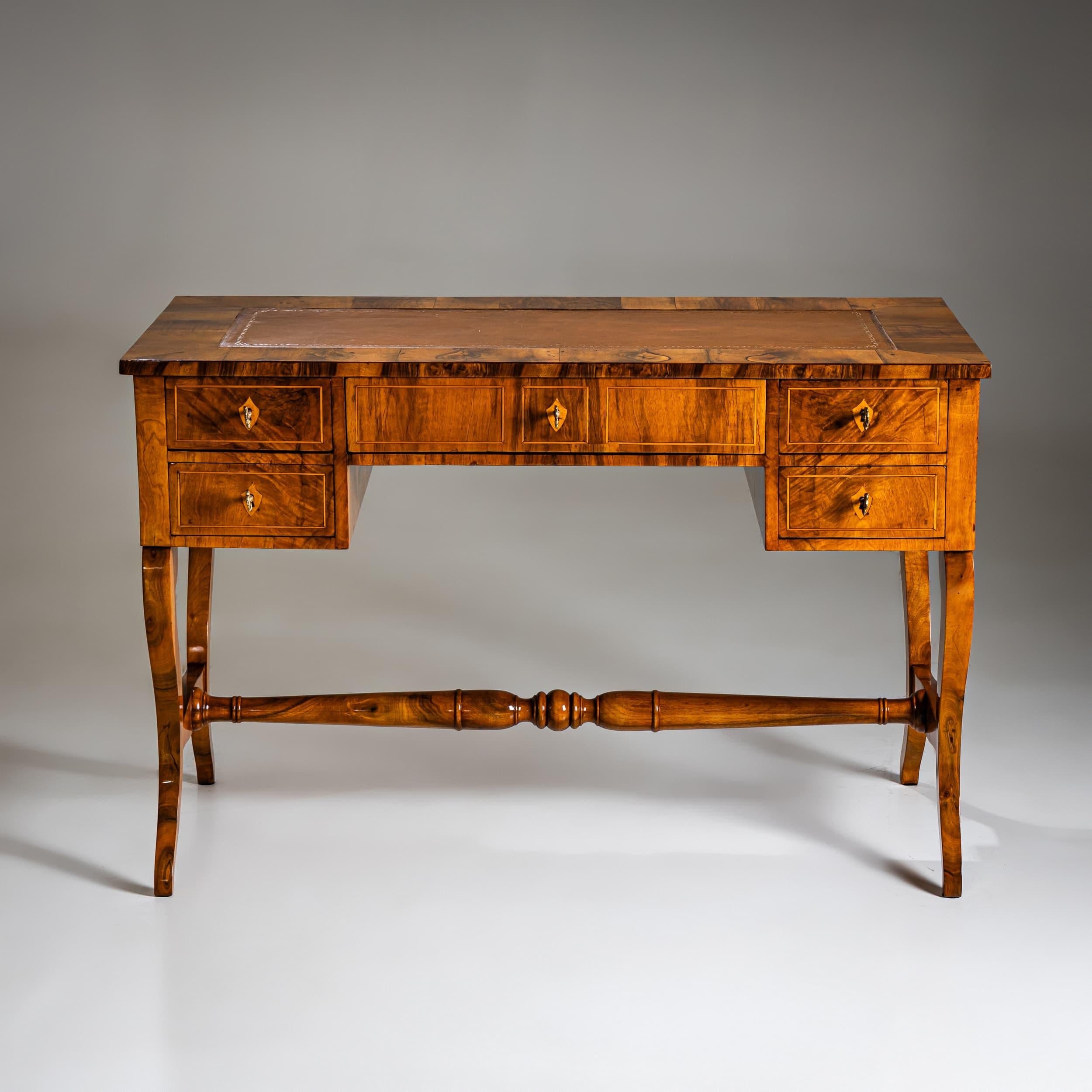 Biedermeier writing desk in walnut, crafted around 1830. Featuring five drawers framed with fine thread inlays and adorned with escutcheons. The gracefully curved S-shaped legs are joined by a turned stretcher. The writing surface is covered in
