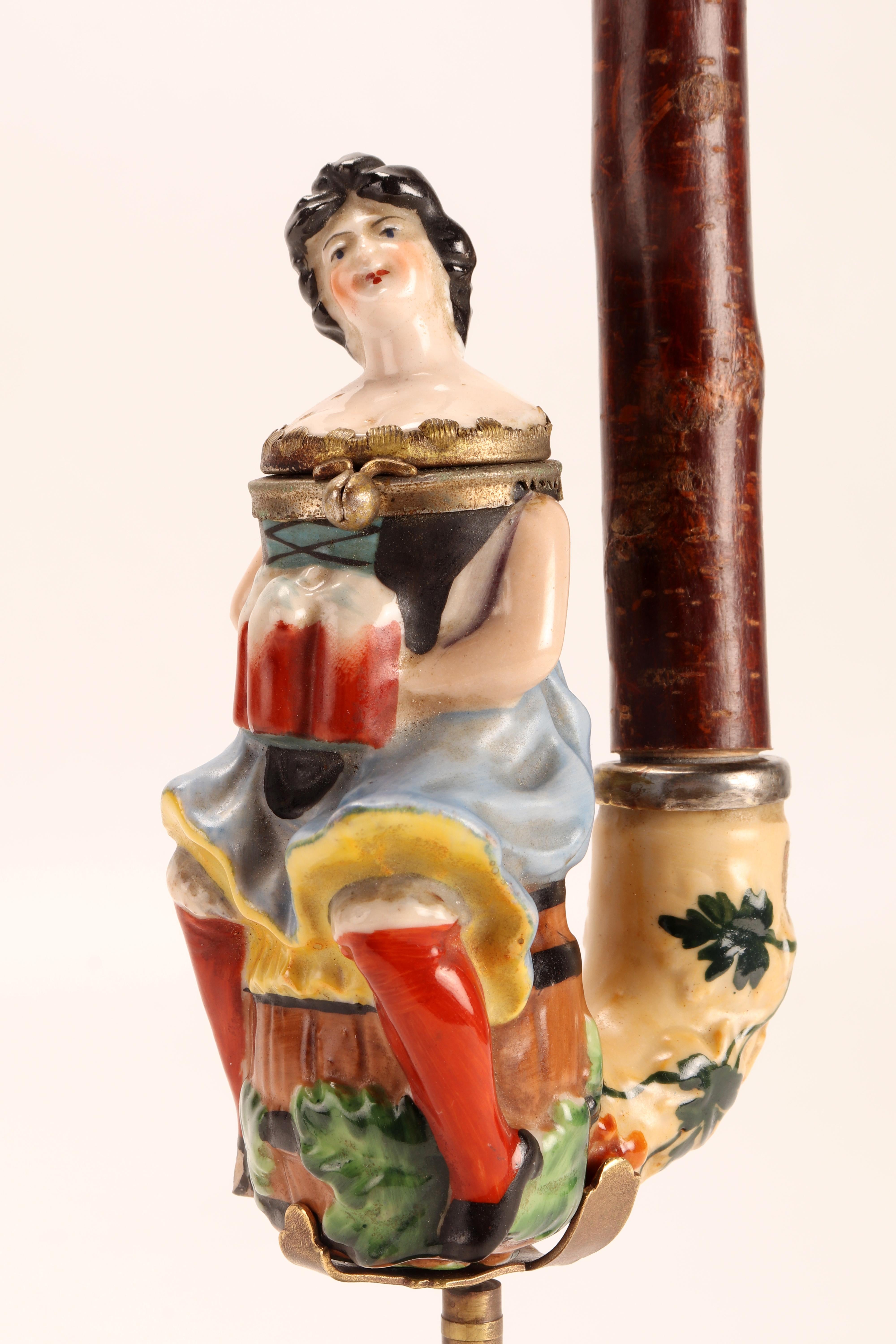 Painted porcelain pipe, with opening stove, depicting a waitress, sitting on a beer barrel, holding three mugs full of beer, ready to be served. The amber mouthpiece is connected to the stove with a connecting branch. Germany circa 1880. (The stand