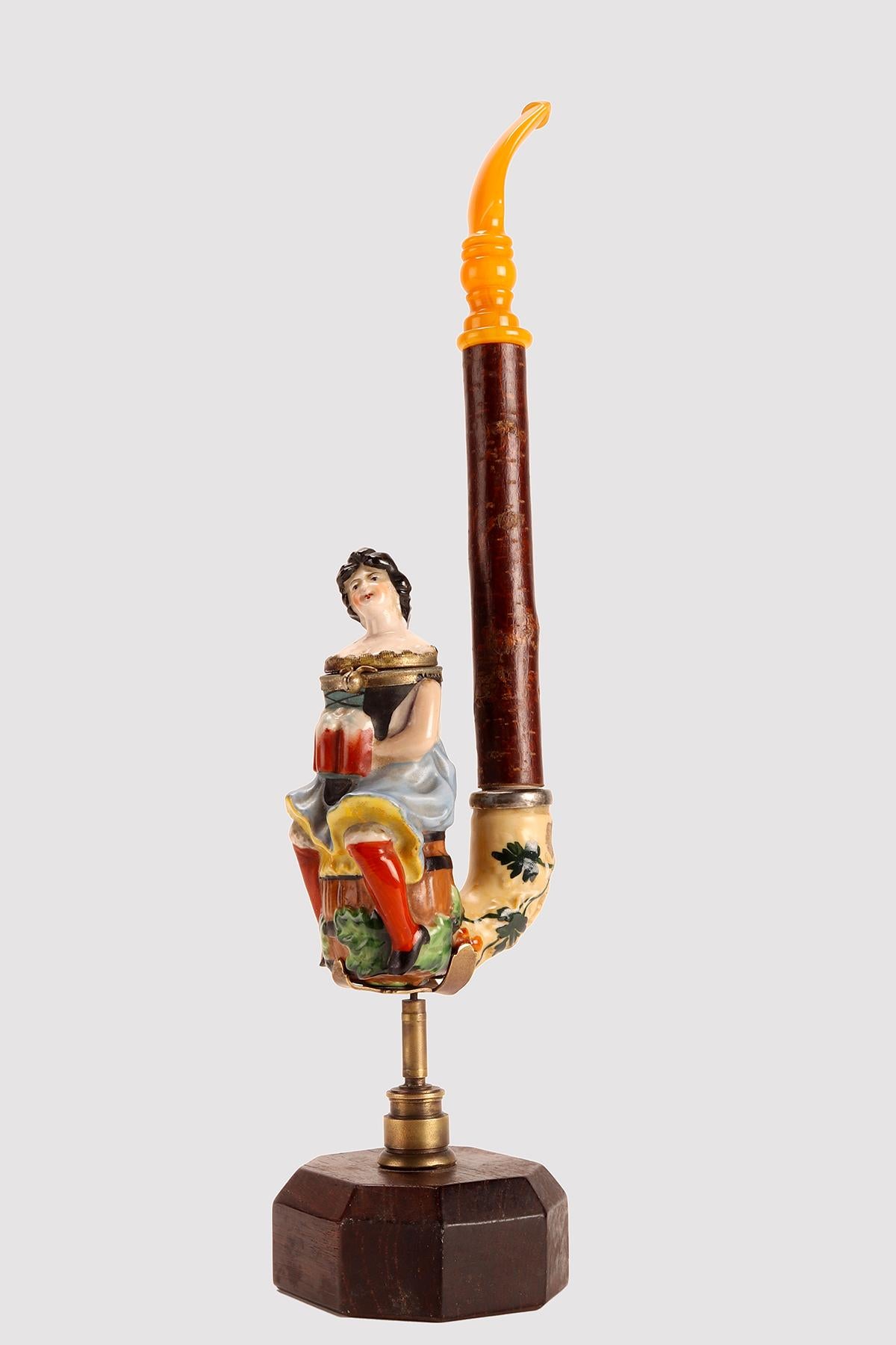 Painted porcelain pipe, with opening stove, depicting a waitress, sitting on a beer barrel, holding three mugs full of beer, ready to be served. The amber mouthpiece is connected to the stove with a connecting branch. Germany circa 1880.  (The stand
