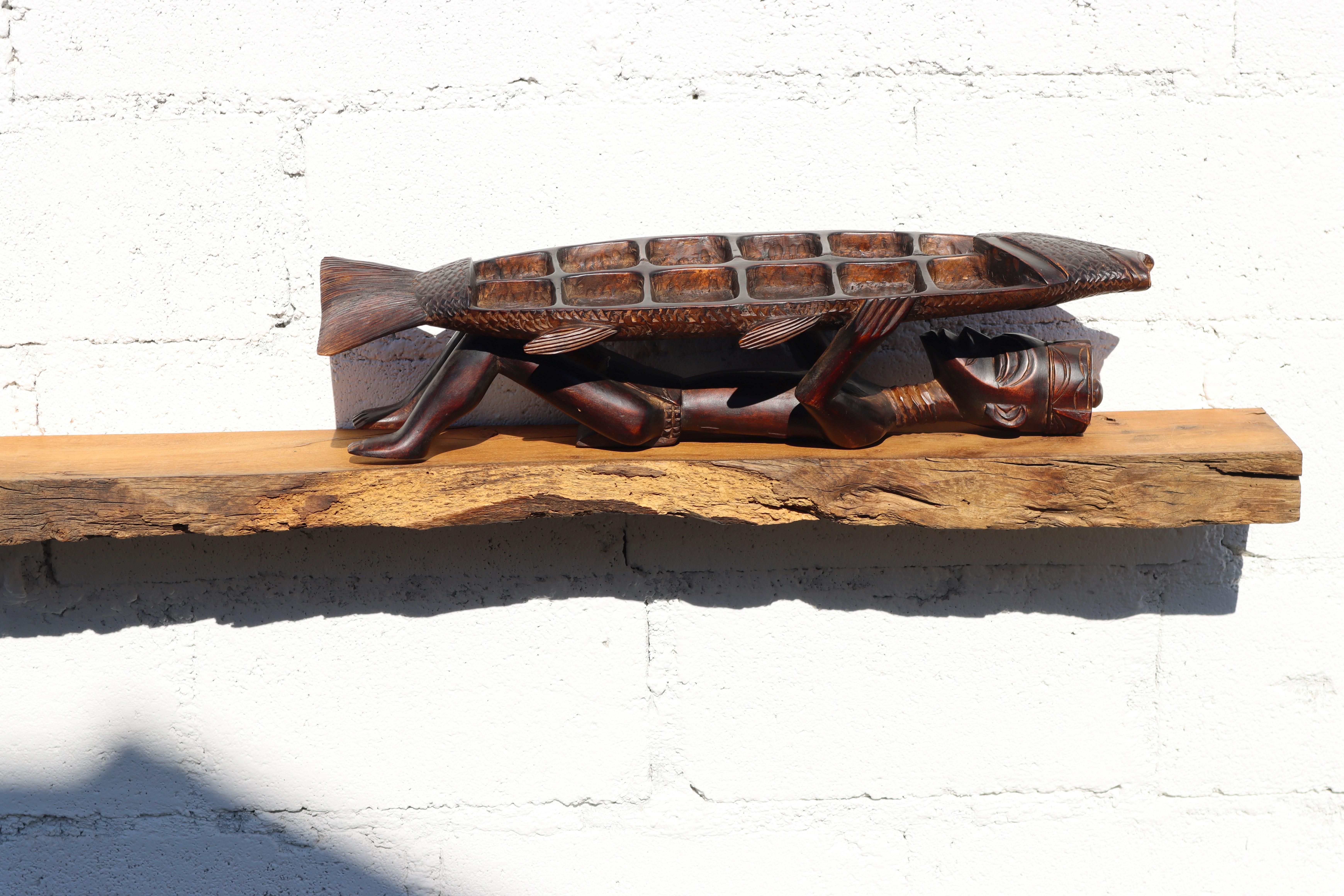 A rare, large African Ebony statue featuring a Tribal Man and Fish from the 1950s. This intricately hand-carved art piece serves as an exquisite table decoration, boasting 12 compartments with a lid ,that does not close precisely, yet this does not