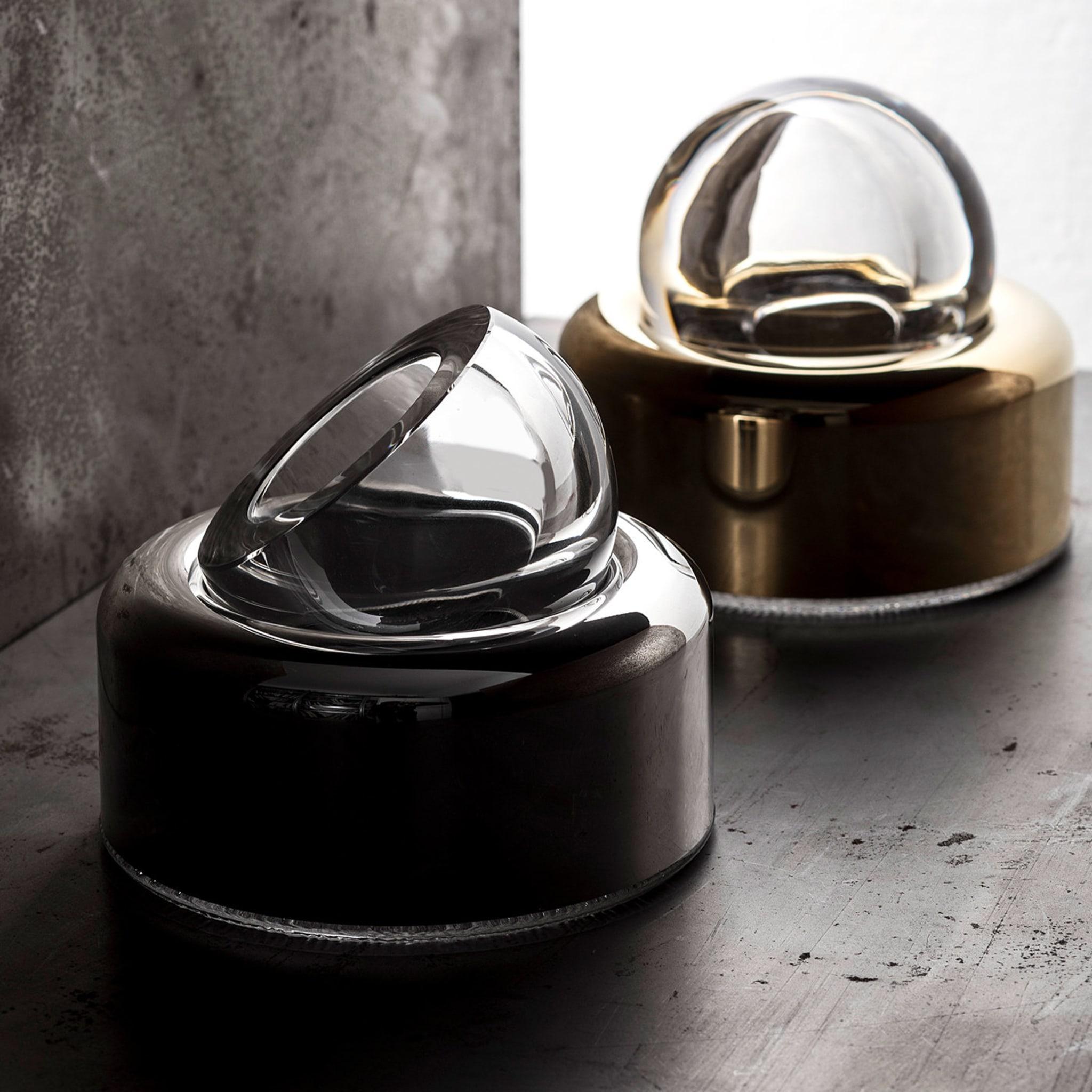 Designed in 1963 by Joe Colombo, this ashtray is part of the Biglia Collection, whose design is centered on the perfect volume of a marble sphere. The top part, made of transparent crystal, rests on a metal base with a gold plating that adds a