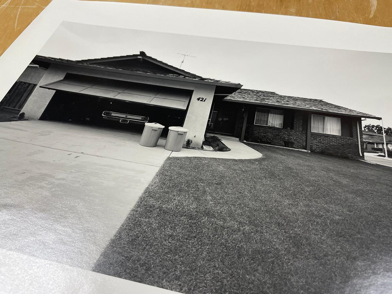 Untitled (Driveway) by Bill Owens presents a tranquil scene. A long driveway leads up to a house in a suburban neighborhood. Two trash cans sit in front of the garage. The garage door is ajar, revealing the tail lights of a parked car. 

 11 x 14