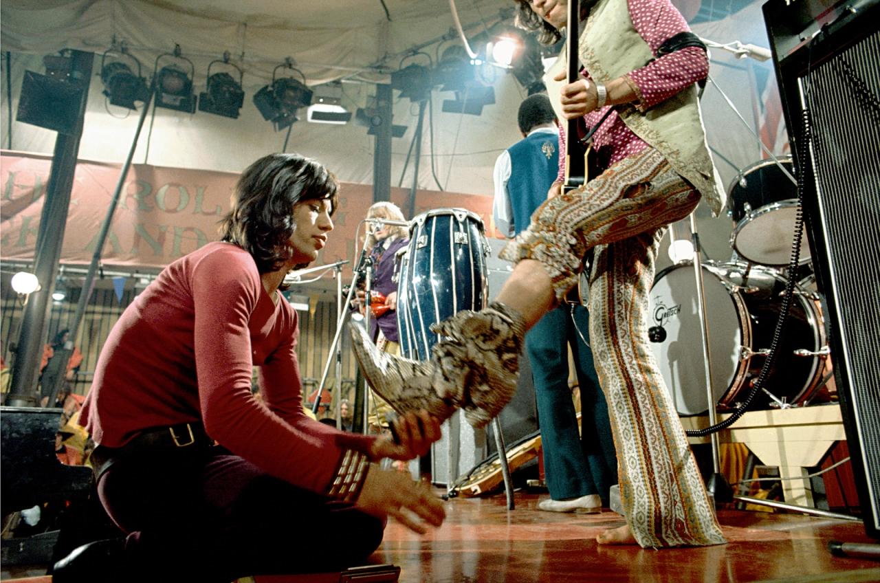 "Mick Giving Keith The Boot", signierter Druck in limitierter Auflage vom Rolling Stones-Bassisten Bill Wyman. 

Mick Jagger hilft Keith Richards mit seinen Schlangenlederstiefeln am zweiten Tag der Dreharbeiten zum Rock and Roll Circus, 11.