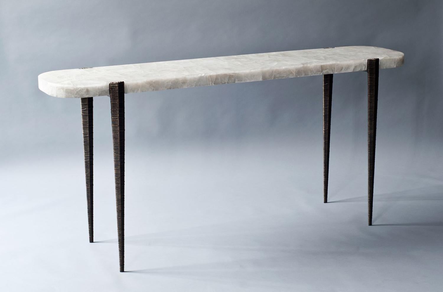 The bind console by DeMuro Das features a stone top in leathered white quartz. The top is supported by elegantly tapered and textured hand-cast legs in solid antique bronze.
