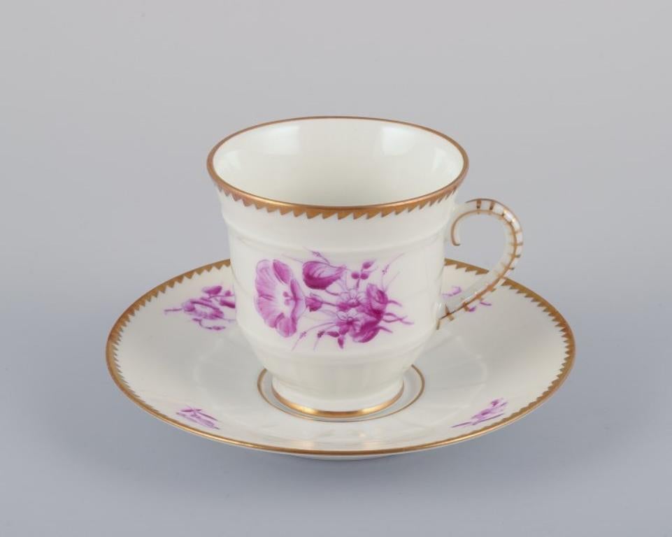 Bing & Grøndahl, Denmark. 
A set of six demitasse cups with saucers decorated with floral motifs in purple and gold trim. Hand-painted.
Approximately from the 1920s.
Marked.
In perfect condition. Appears unused.
First factory quality.
Cup: Height