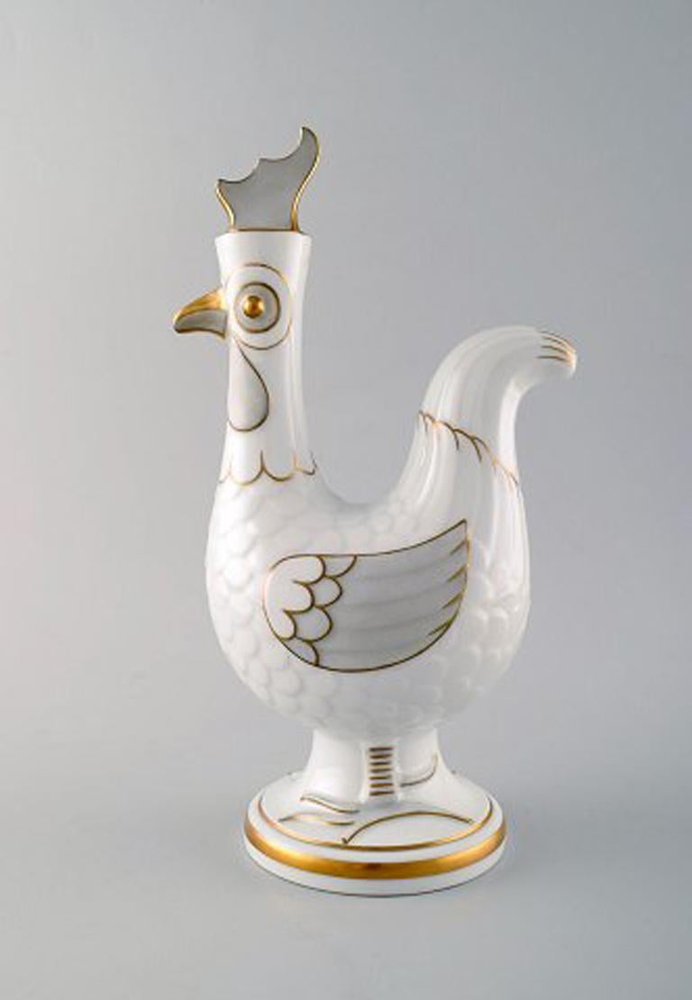 Bing & Grondahl/ B&G, Denmark. 
Jug of porcelain in the form of rooster, stopper with rooster comb. 
12 accompanying porcelain mugs with gold-painted details. 
The jug measures: 28 x 16 cm. The cups measure: 6 x 5 cm.
In very good