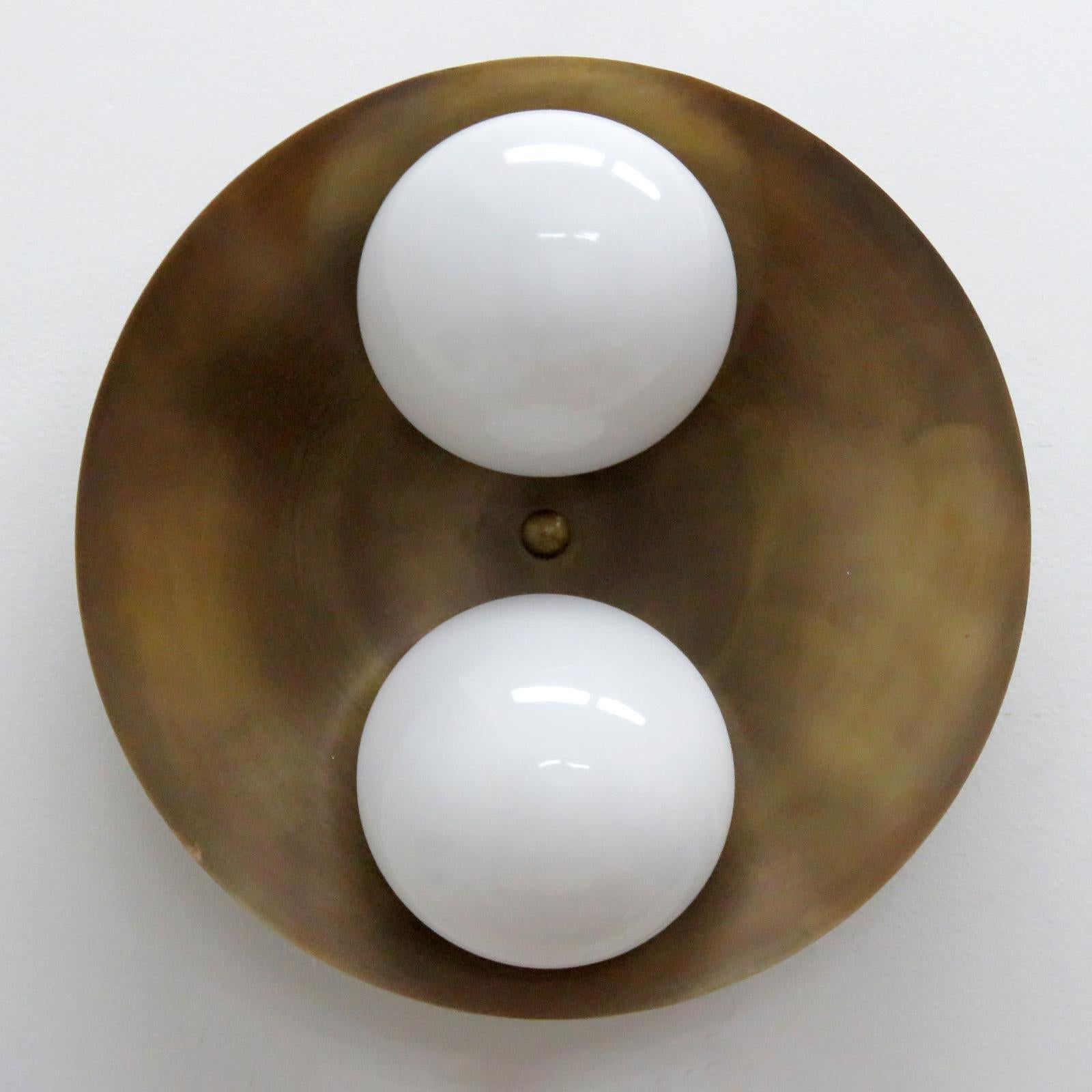 Elégante applique ou plafonnier Binova conçue par Gallery L7, fabriquée et finie à la main à Los Angeles en laiton américain, avec deux abat-jours en verre opalin sur un disque en laiton brut vieilli. Deux douilles E12 par luminaire, puissance