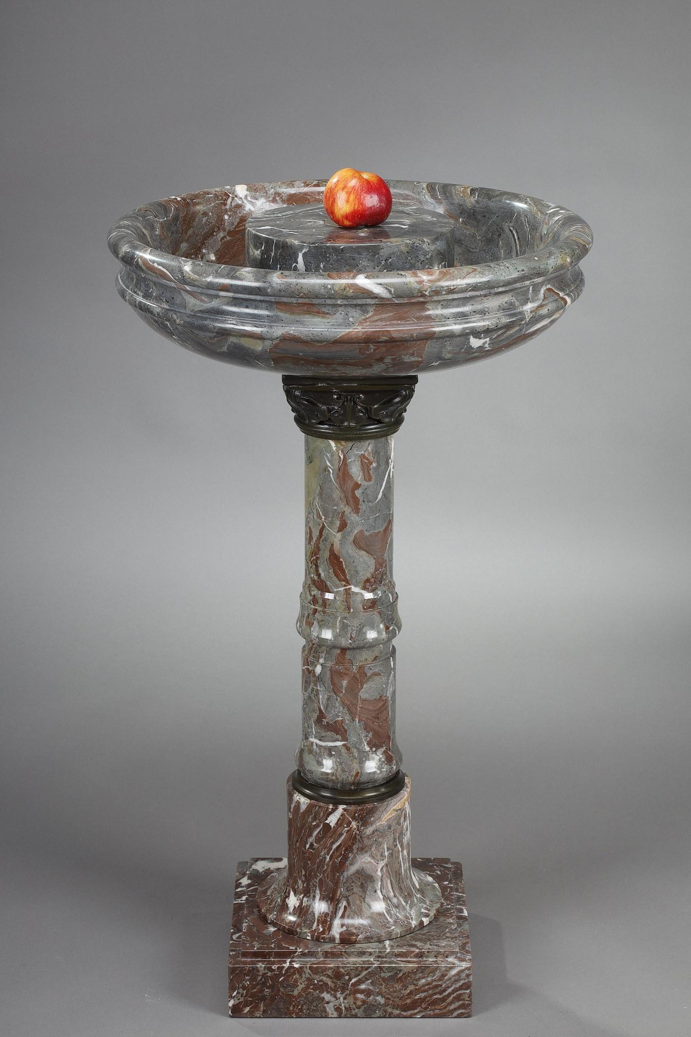 A grey and red marble birdbath. It rests on a shaft in the form of a ringed column. The capital of water leaves is in patinated bronze. The round basin with its moulded lip has a central pedestal that can accommodate a sculpture. The whole rests on