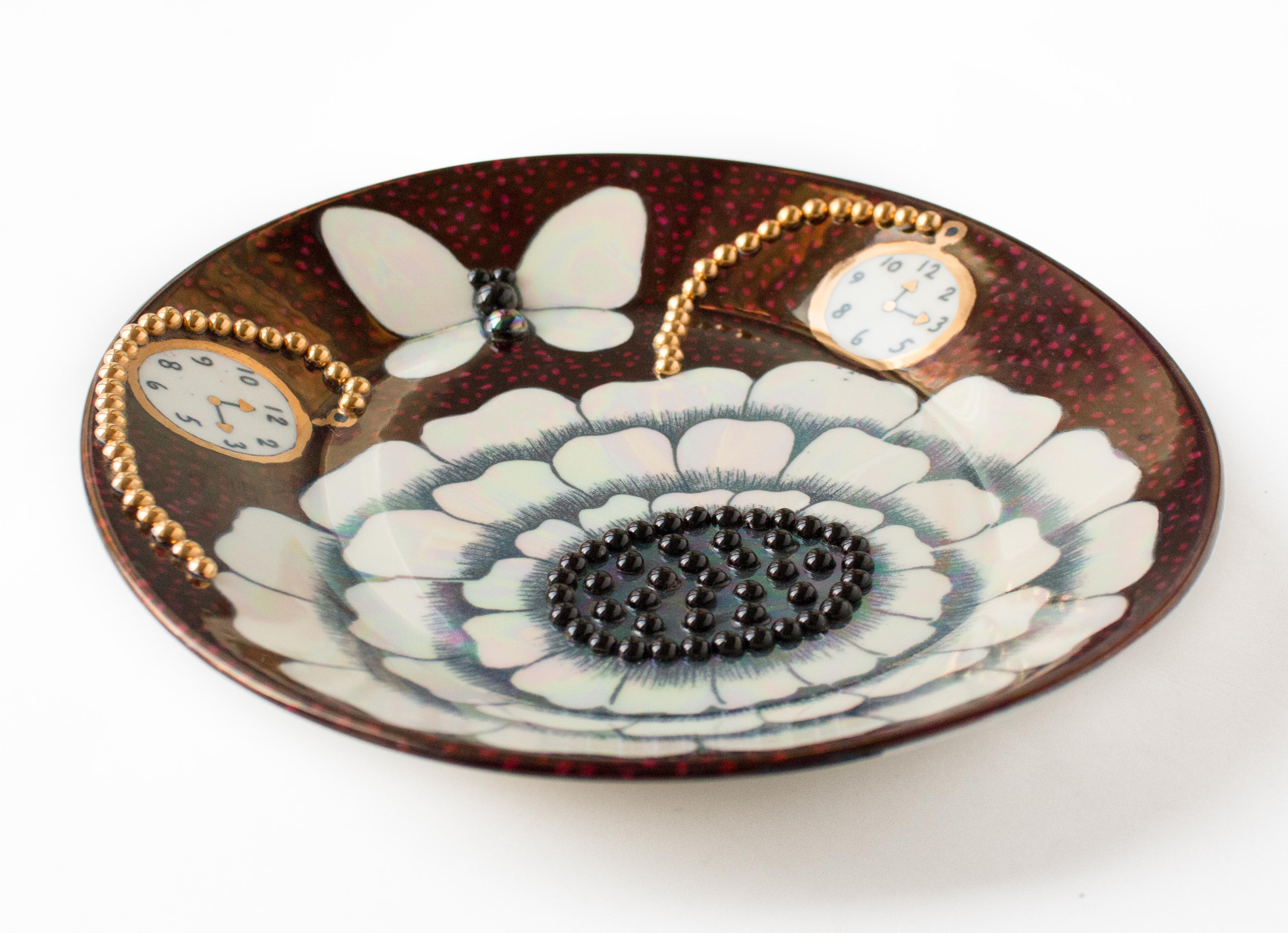 This beautifully glazed stoneware plate, measuring 25 x 23 cm, is a stunning example of Birger Kaipiainen’s artistry, featuring intricate bead details and a lustrous glaze. The decoration includes a delicate flower, a butterfly, and a pocket watch