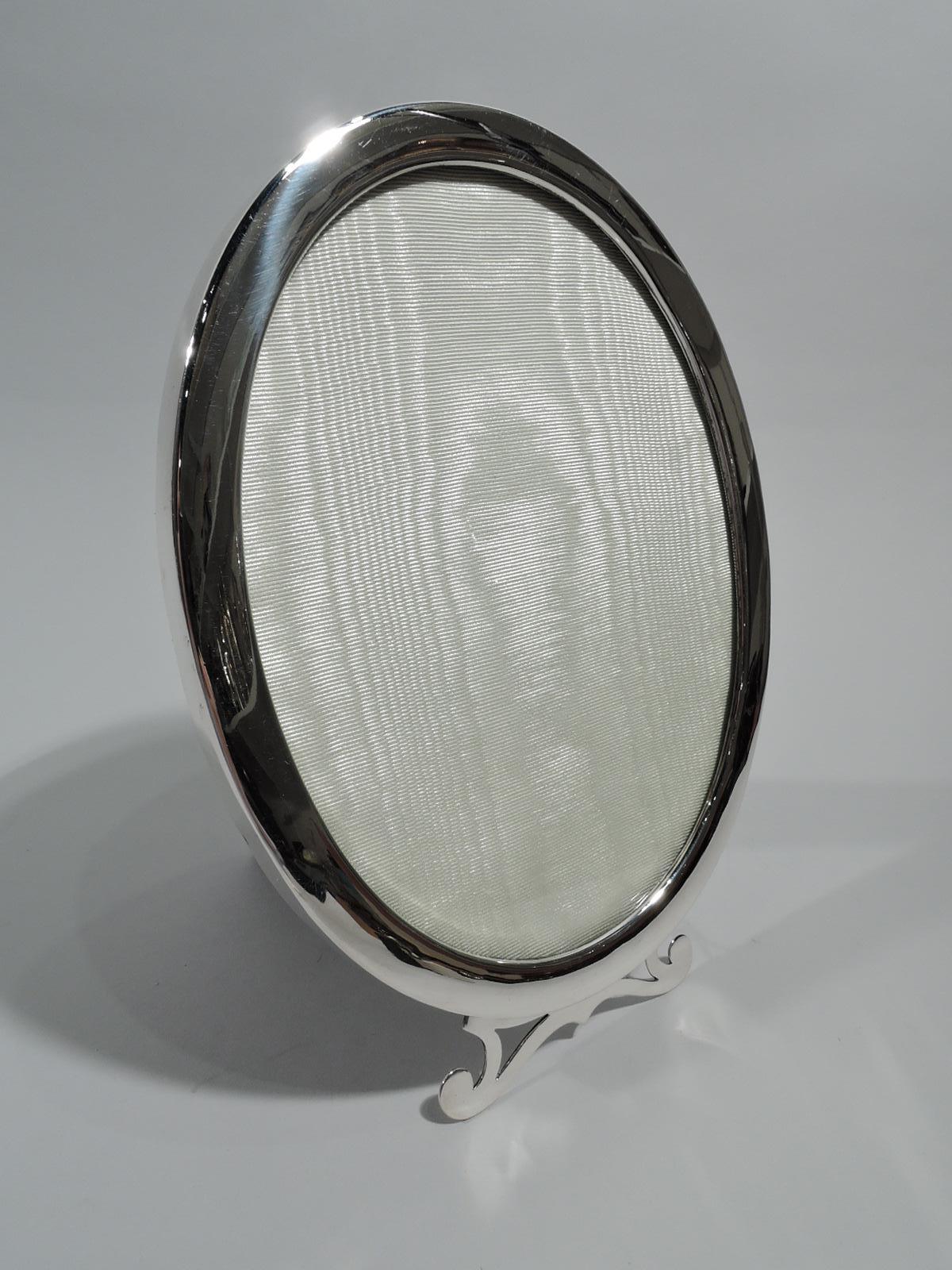 Modern classical sterling silver picture frame. Made by Birks in Canada, ca 1950. Oval window in plain surround on open scrolled support. With glass, silk lining, and velvet back and hinged and chained easel support. Marked “Birks / Sterling”.