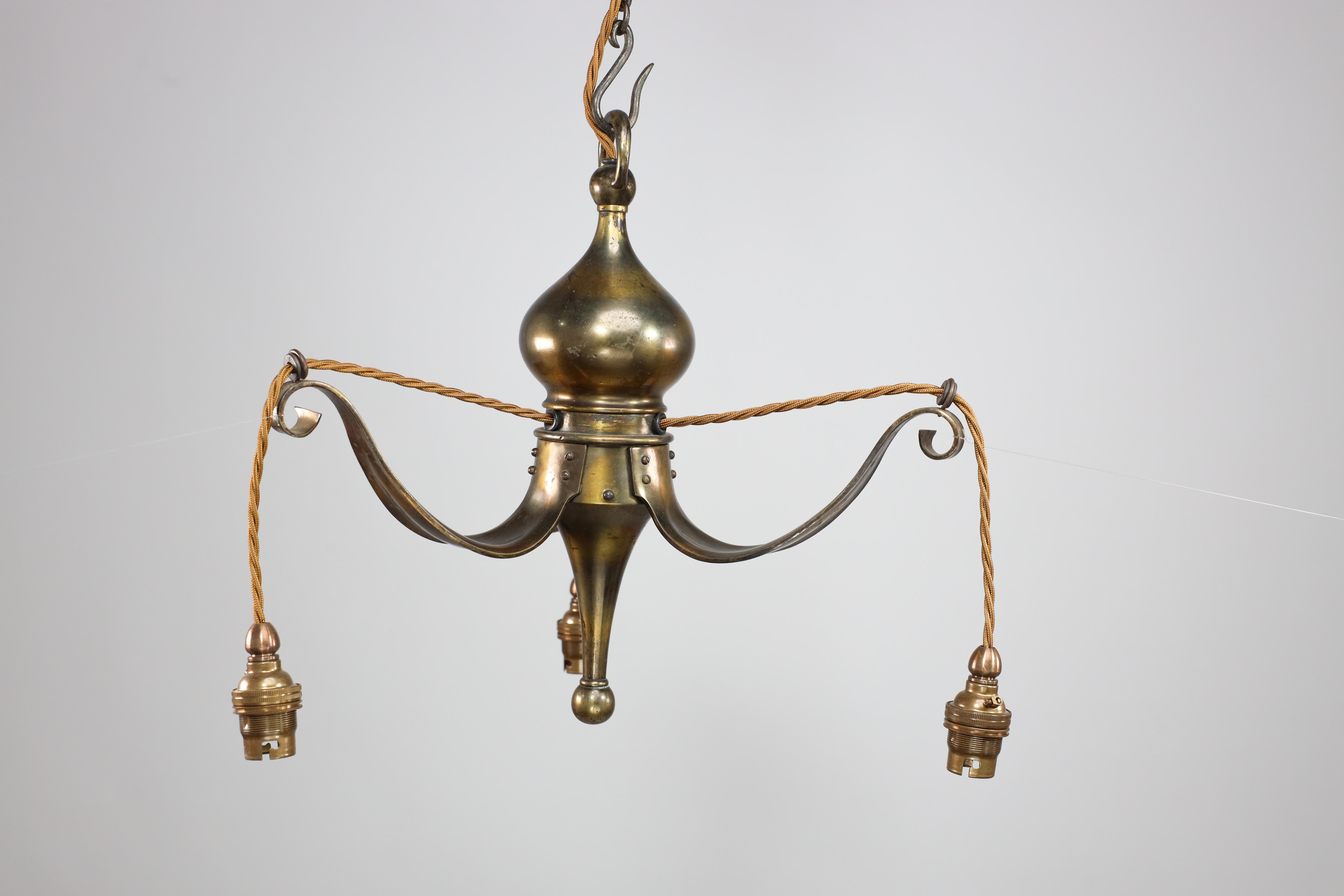 Birmingham Guild of Handicraft. An Arts and Crafts brass ceiling light or chandelier with three curved arms all hand riveted to the main bulbous body with an elongated turned finial to the base.