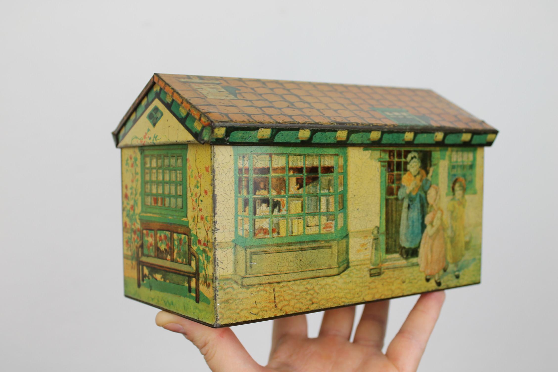 Biscuit tin house. 
A vintage biscuit tin - cookies tin in the shape of a house. 
The tile roof of this tin box can be opened. 
Around you have different scenes: 
little grocery shop, mama waving the kids out who will go to school, a bench against