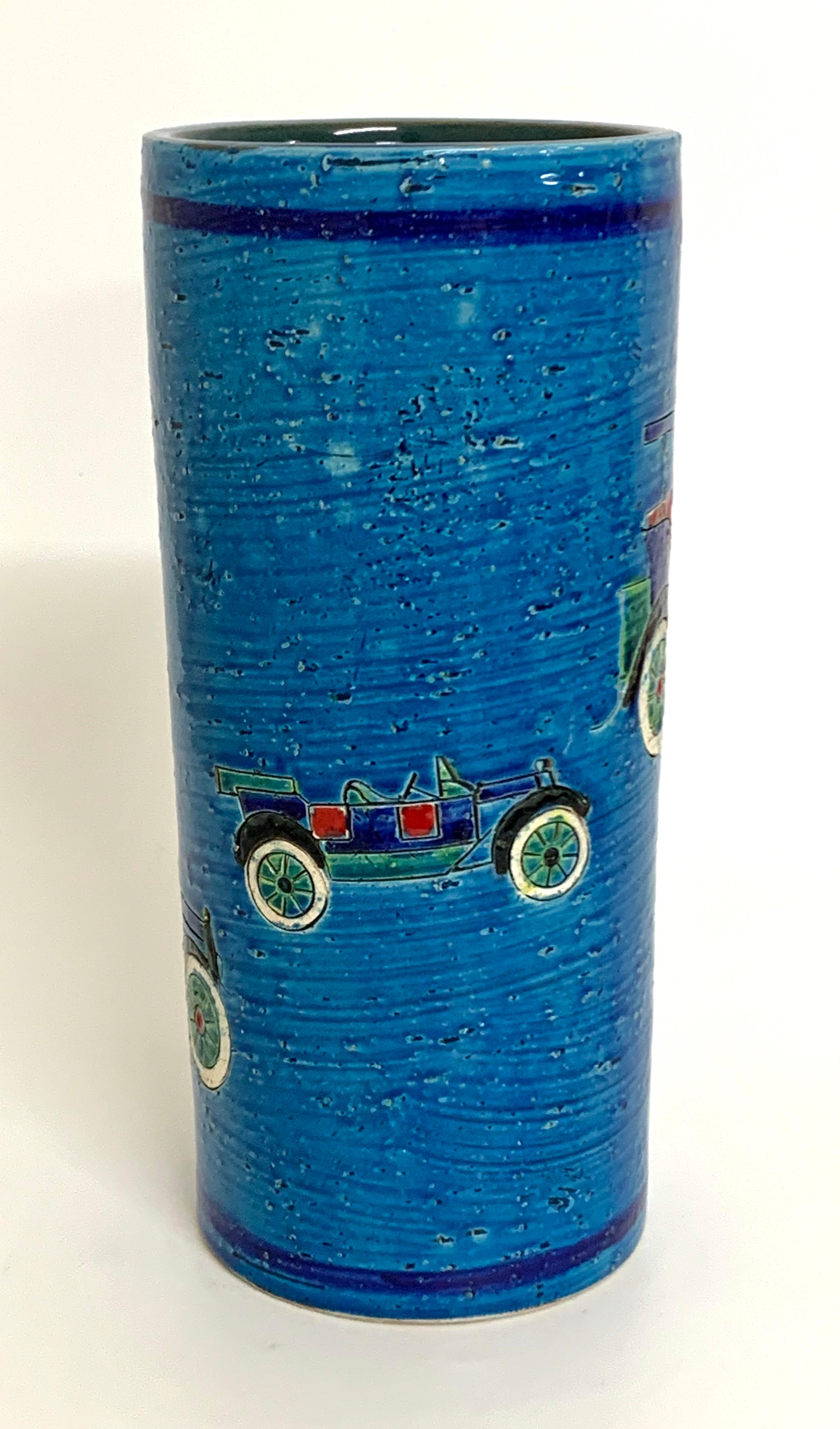 A bright blue cylinder vase decorated with 3 different vintage cars. The cars outlines are deeply incised and coloured with various coloured glazes. It retains part of its Rosenthal Netter paper label to the base along with its pattern number and