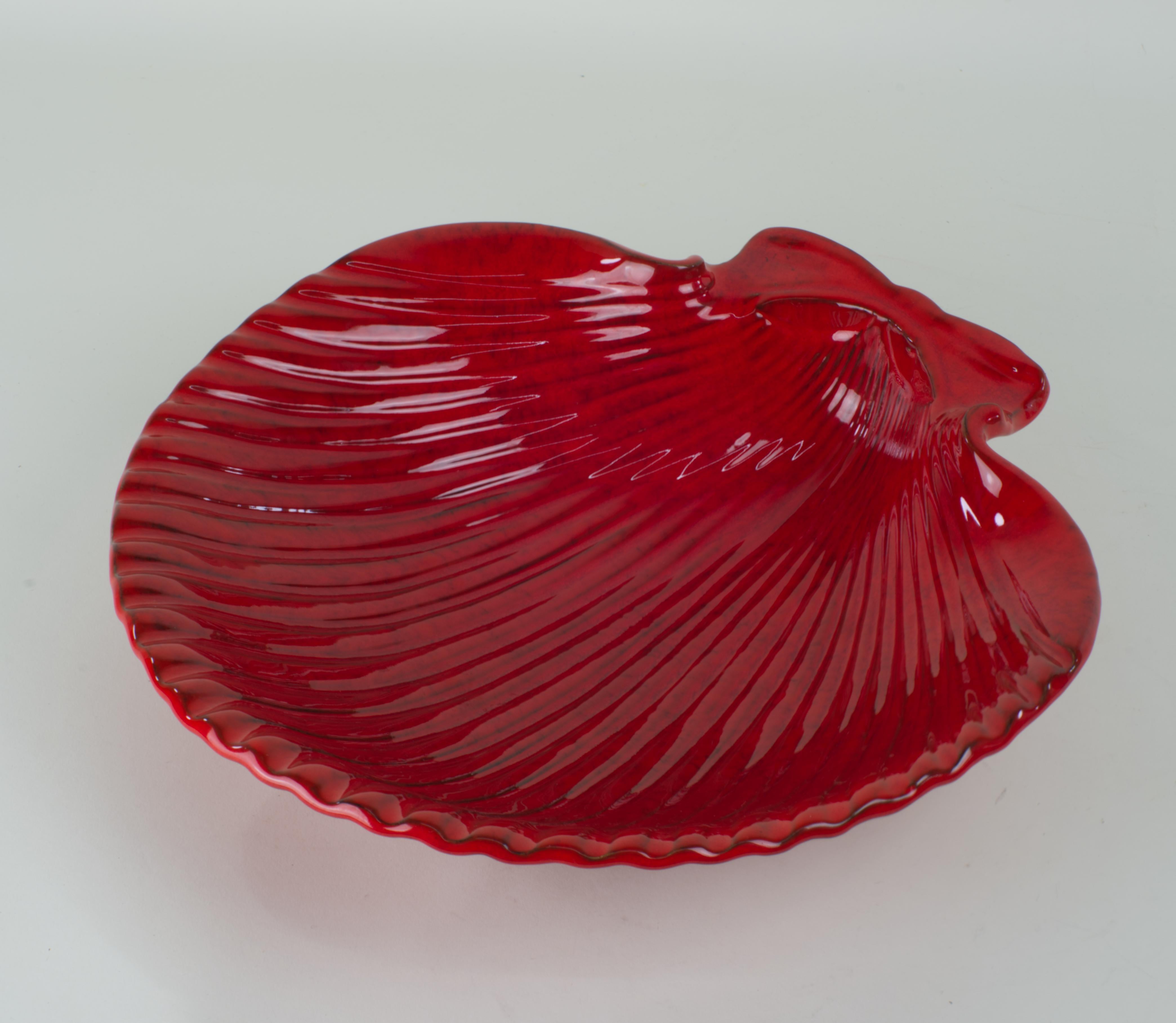 
Shell-shaped ceramic bowl is glazed in red with complex variations in glaze color accentuating the intricate shape of the bowl. 

The bowl is signed on the bottom with 