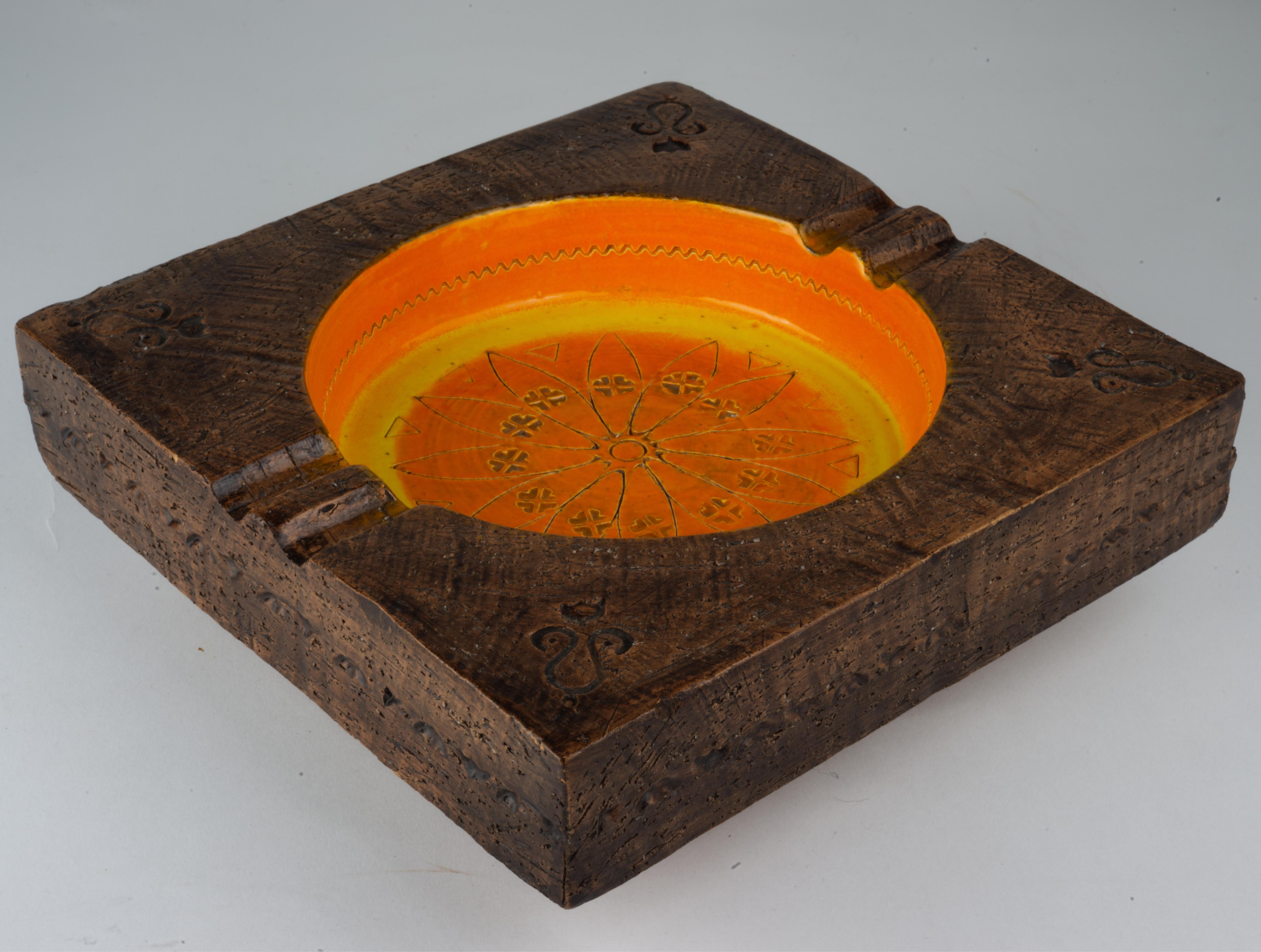  Mid-Century Modern catch all or ashtray was made by Bitossi for Rosenthal Netter. The square semi-matte brown outer rim encloses the concave circle in the middle that is decorated with bright orange and yellow glaze with incised accents. The piece