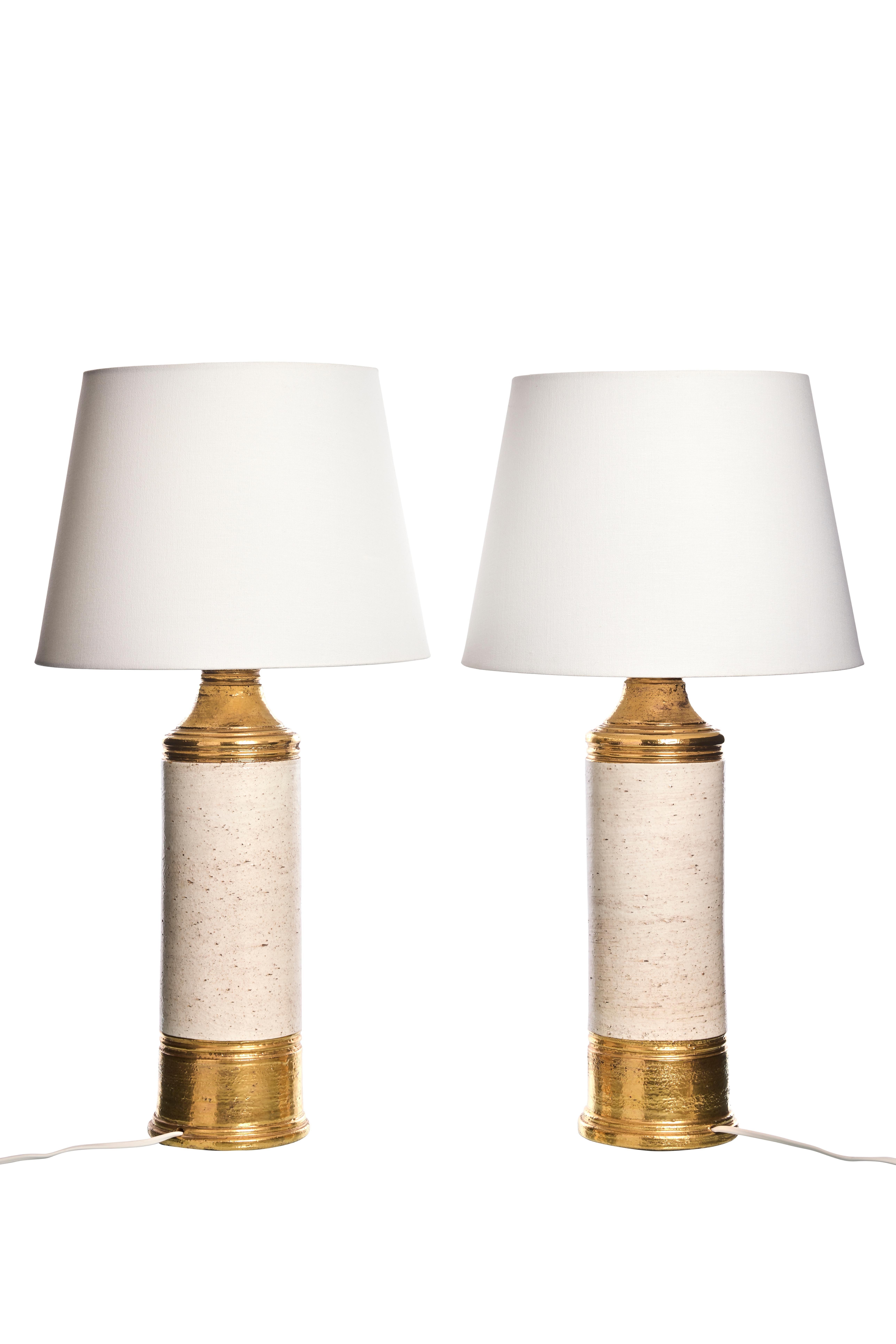 Chaque lampe présente une base cylindrique avec une combinaison frappante de céramique émaillée blanc cassé et de riches accents dorés. La glaçure blanc cassé donne un aspect texturé et naturel qui contraste magnifiquement avec les détails dorés