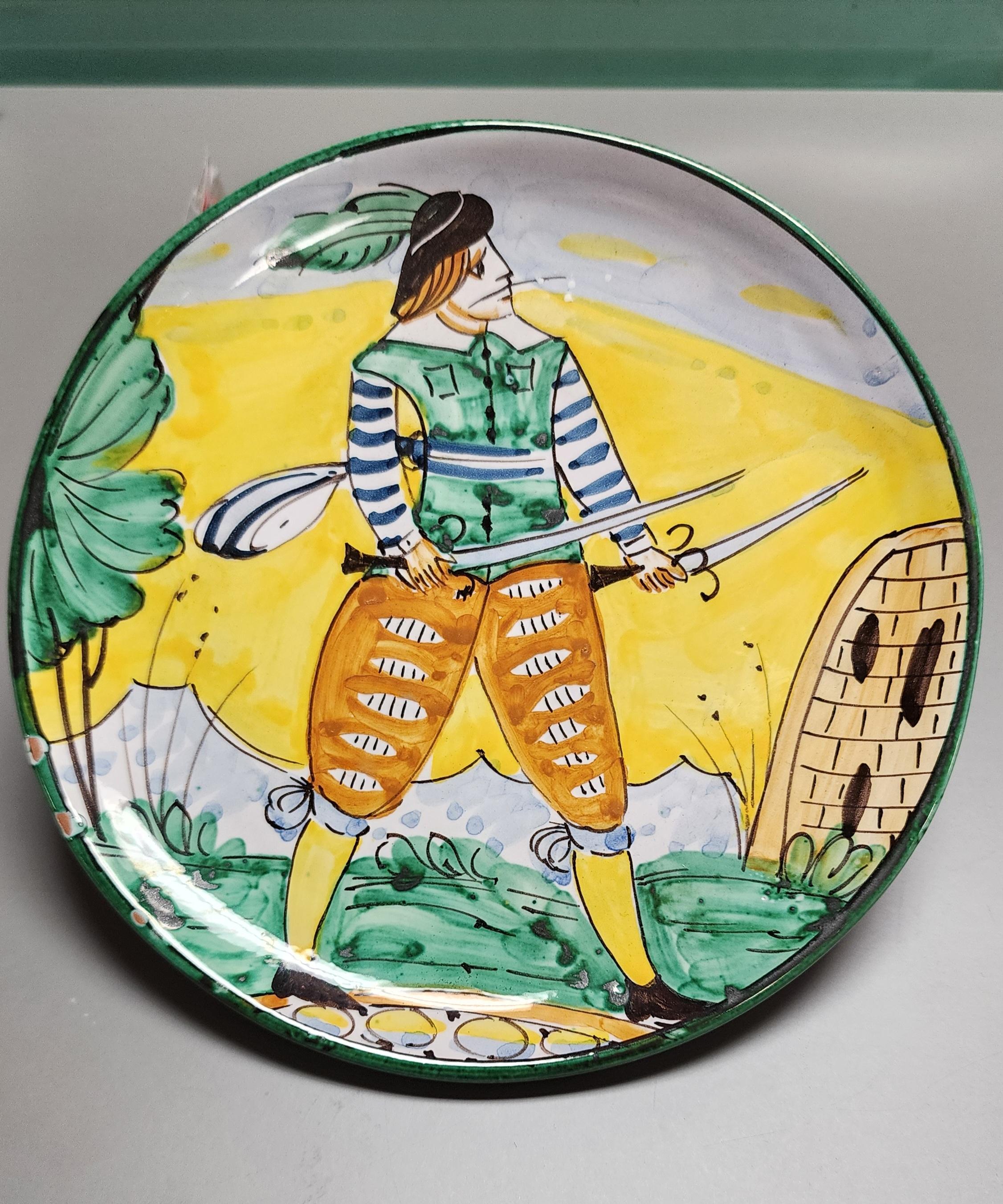 This Italian Pottery plate was made by the famed Bitossi Company. The art is of a soldier in traditional Italian garb wielding two swords. The vivid colors of the clothing along with the vibrant yellow background are typical of this design. 
The