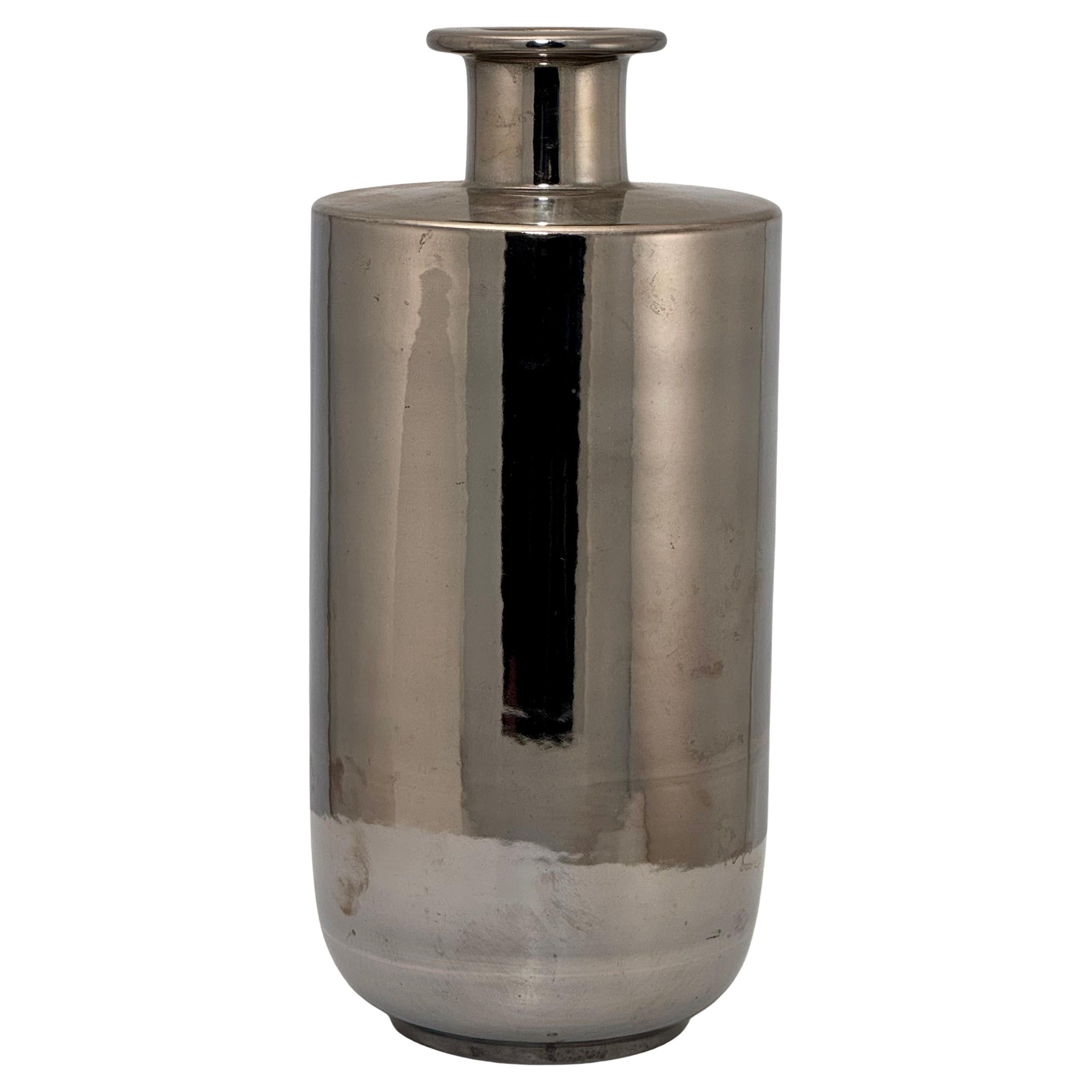Ce vase est cylindrique, avec une base inversée et une embouchure en forme de bougeoir. Il est recouvert d'une glaçure chromée/argentée qui lui donne l'apparence du métal. Il porte l'étiquette d'origine en dessous.