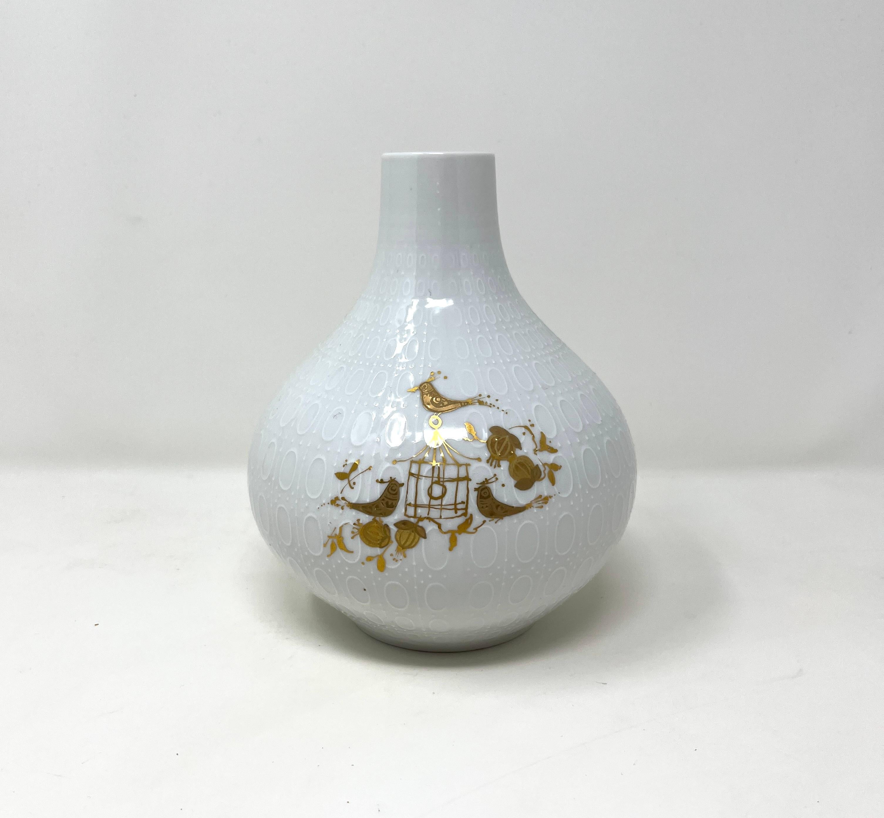 Vase Bjorn Wiinblad pour Rosenthal en forme de gourde en porcelaine avec relief clouté et ovale. Peint à la main à la feuille d'or, un personnage enjoué se pavane parmi les citrouilles, un motif enfantin typique de l'art de Wiinblad.

(L'or est