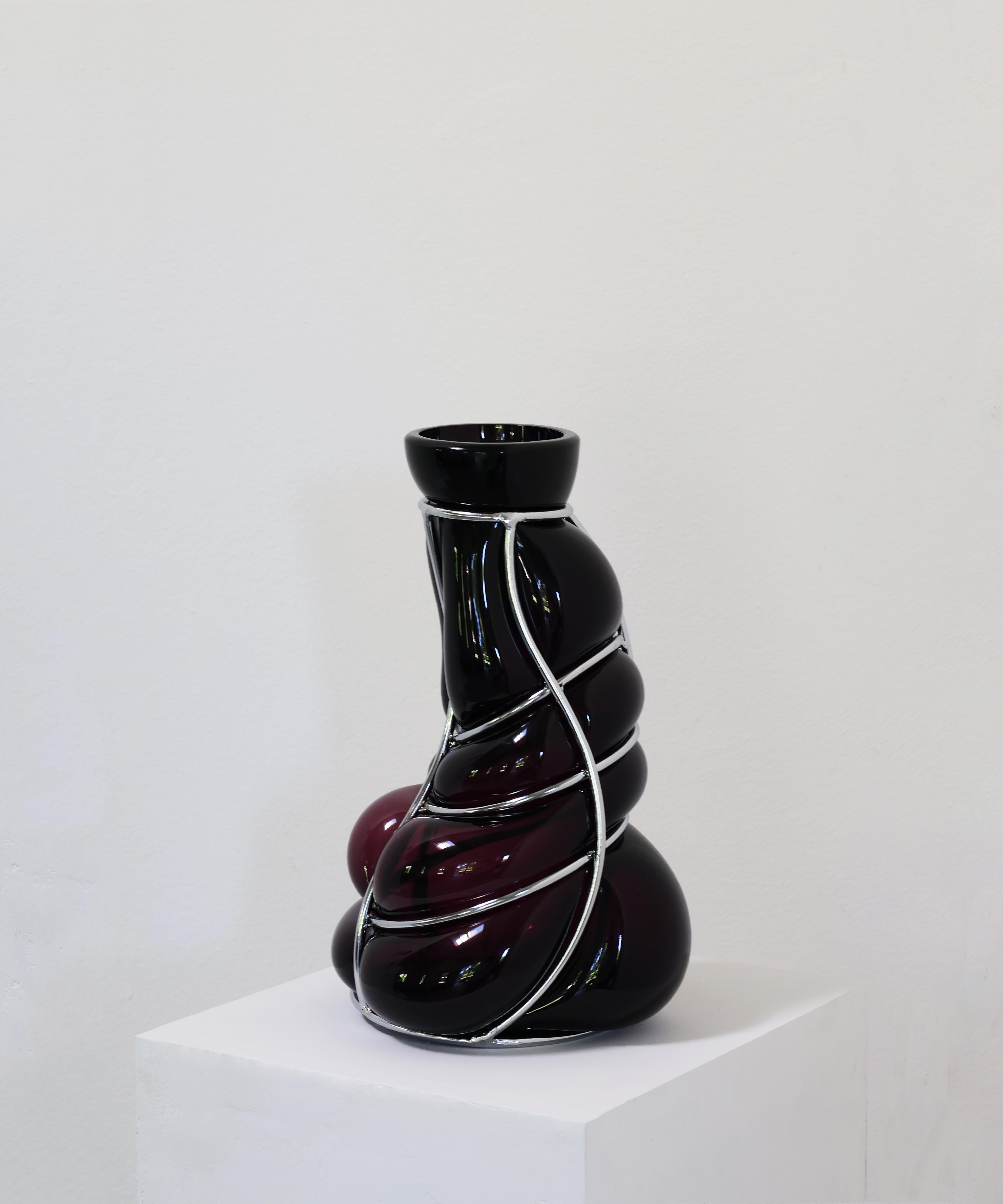 Vaso scultoreo Babà in ametista nera di Alice Crepaldi
Un pezzo unico.
Dimensioni: Ø 20 x H. 36 cm.
Materiali: Vetro di Murano e metallo.

Adornato da uno stelo leggermente più corto ma elegante, questo vaso ingabbiato utilizza la tecnica dello
