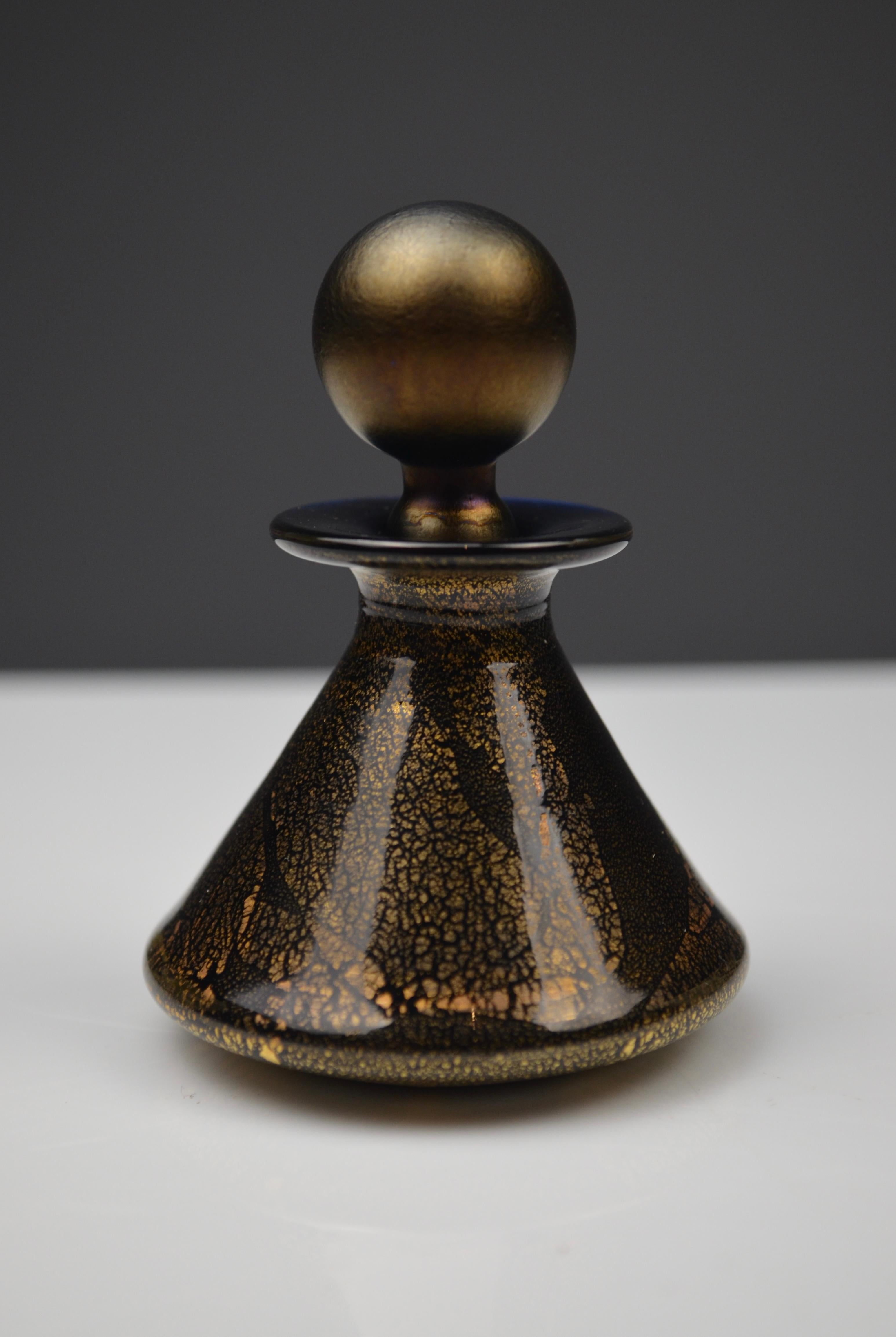 Elegante bottiglia di profumo in vetro soffiato firmata Isle of Wight Studio Glass.
La sua forma conica è impreziosita da un ricco design nero e oro con effetti crepati e metallici, che creano un raffinato gioco di luci. Il tappo sferico satinato