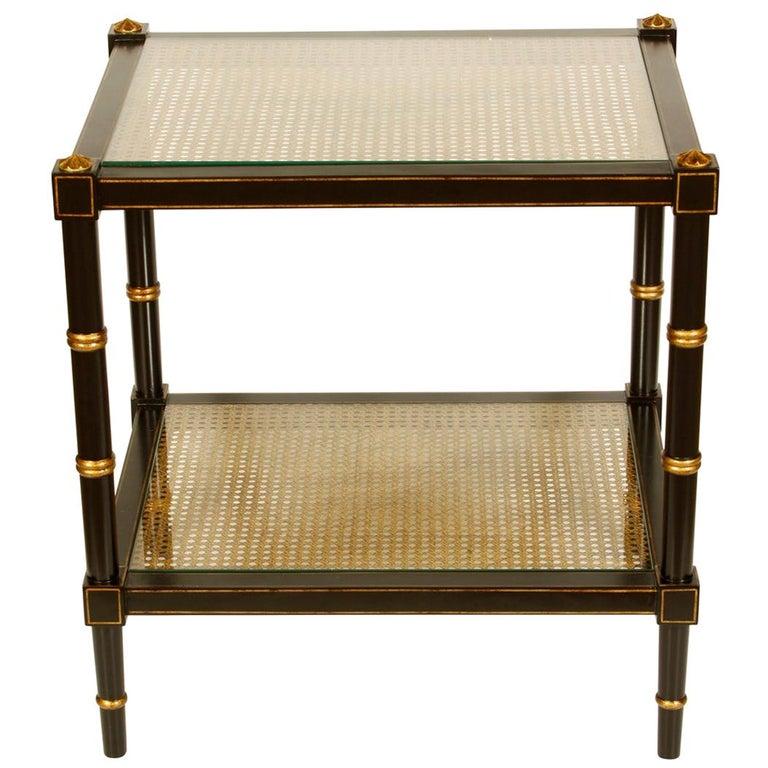 A handsome two-tiered table in black with gold accents featuring two caned glass shelves. 
