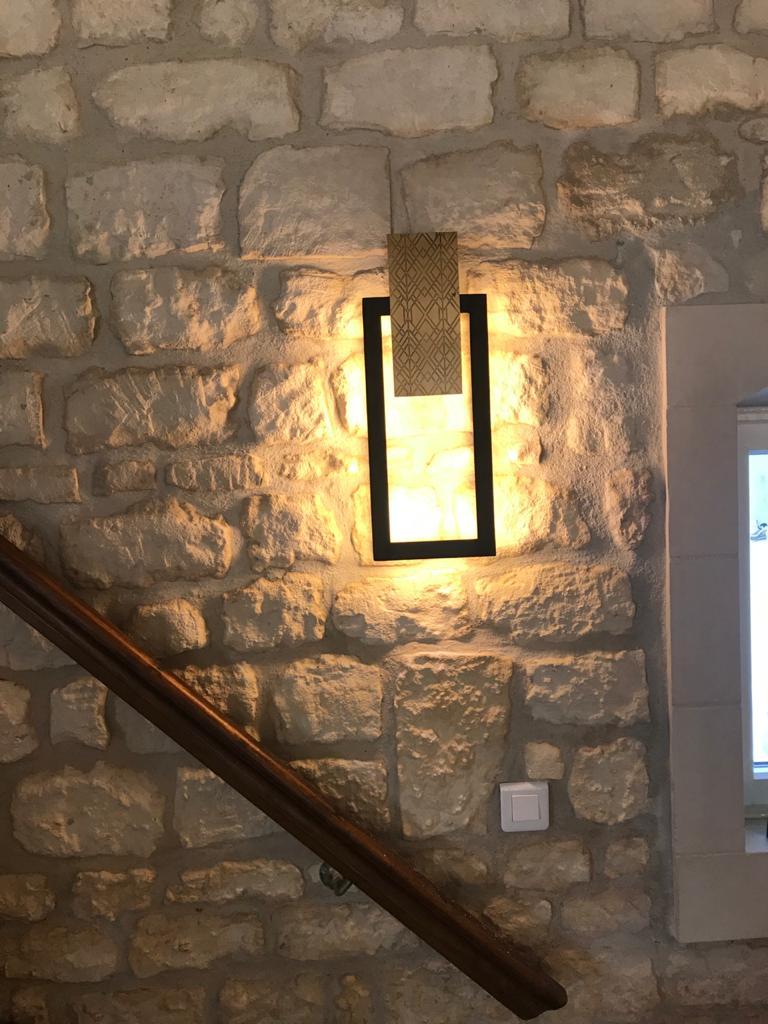 El aplique negro y dorado Frank 3 presenta un juego de líneas limpias y elementos en capas. Una placa central de latón grabado, ligeramente desplazada de la pared, está enmarcada por una estructura rectangular de acero negro, que crea un contraste