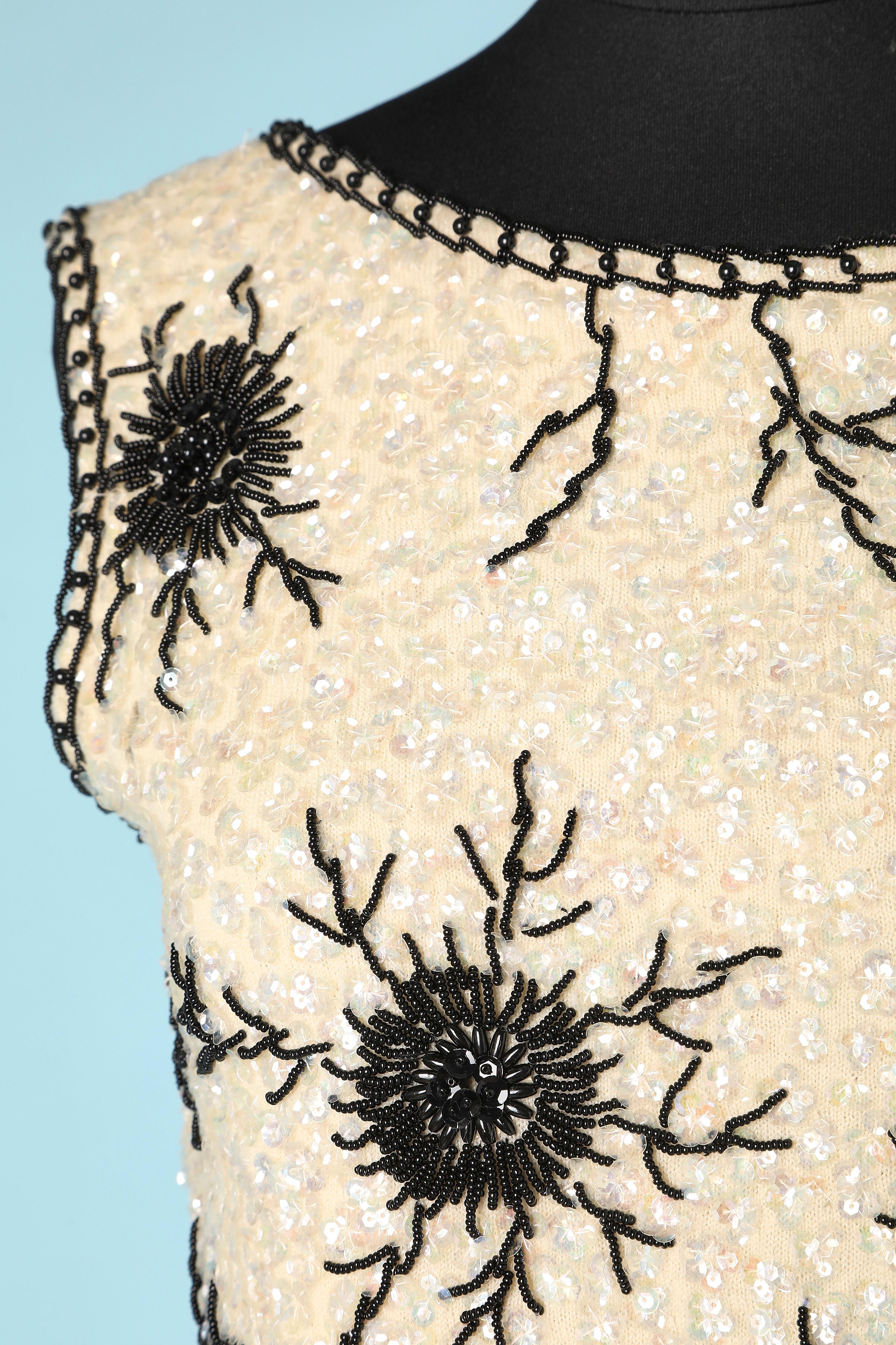Top senza maniche con paillettes e perline bianche e nere su base in maglia di lana. Fodera in acetato o rayon. Chiusura con zip e gancio sulla parte centrale della schiena. Spaccato su entrambi i lati con chiusura a gancio.
La taglia S riportata