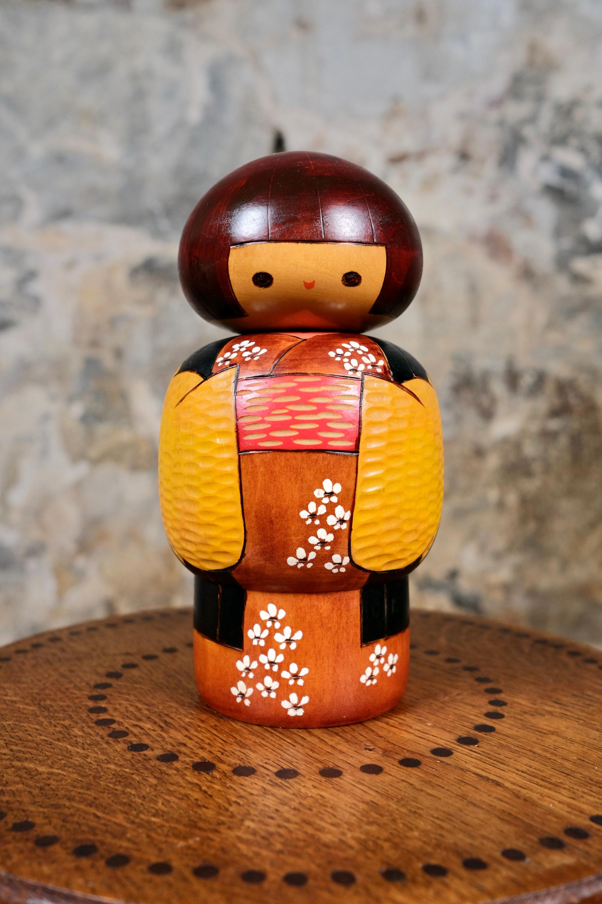 Kokeshi à kimono jaune et noir, des années 1970, par Bitoku. Une kokeshi rare avec un joli visage souriant, des couleurs chaleureuses et un délicat travail de sculpture et peinture. Rare, en très bon état, signée.
H22,5 D12

D'autres kokeshis sont