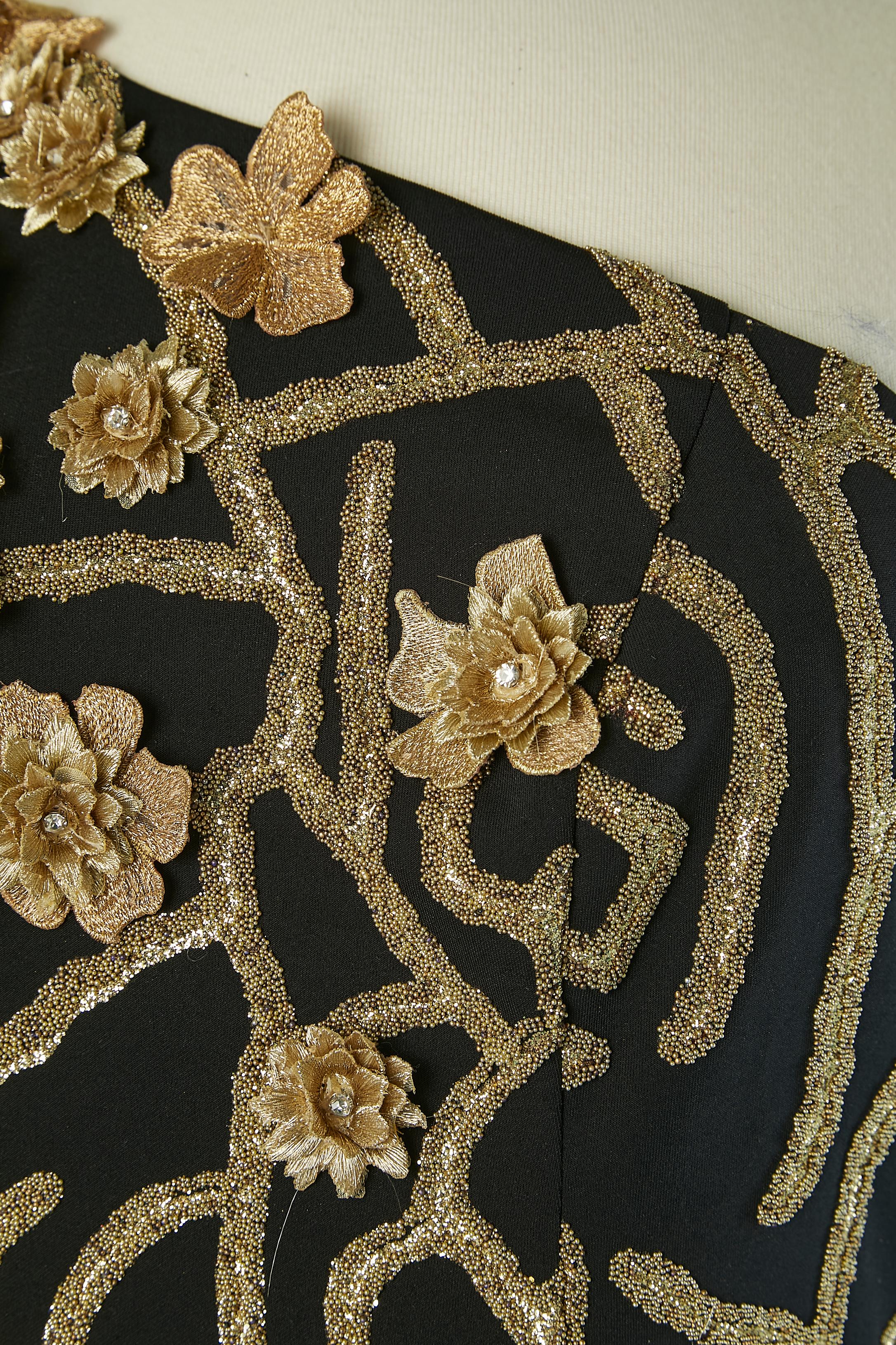 Vestido de noche negro de arrastre asimétrico con adornos dorados de flores, cuentas y pedrería. Tejido principal: poliéster. Cremallera invisible en el lado izquierdo. Inserto de tul transparente en la espalda.
Longitud total en el centro delantero