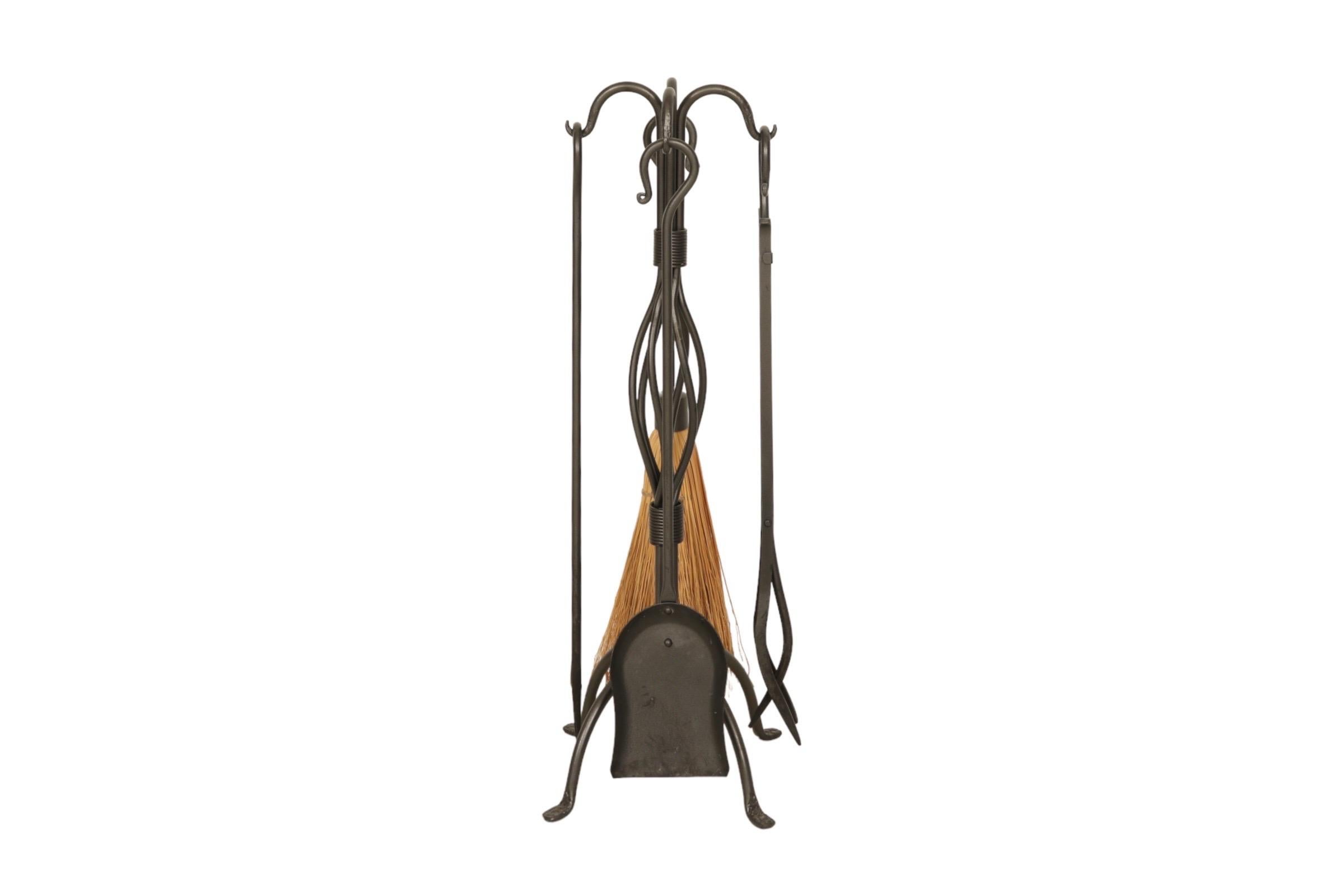 A set of black cast iron basket weave fireplace tools made by Pilgrim. An ash rake, poker, shovel and brush each have a curved hook handle. Tools hang from a cast iron yoke with an intertwined hand forged iron centerpiece supported on four arched