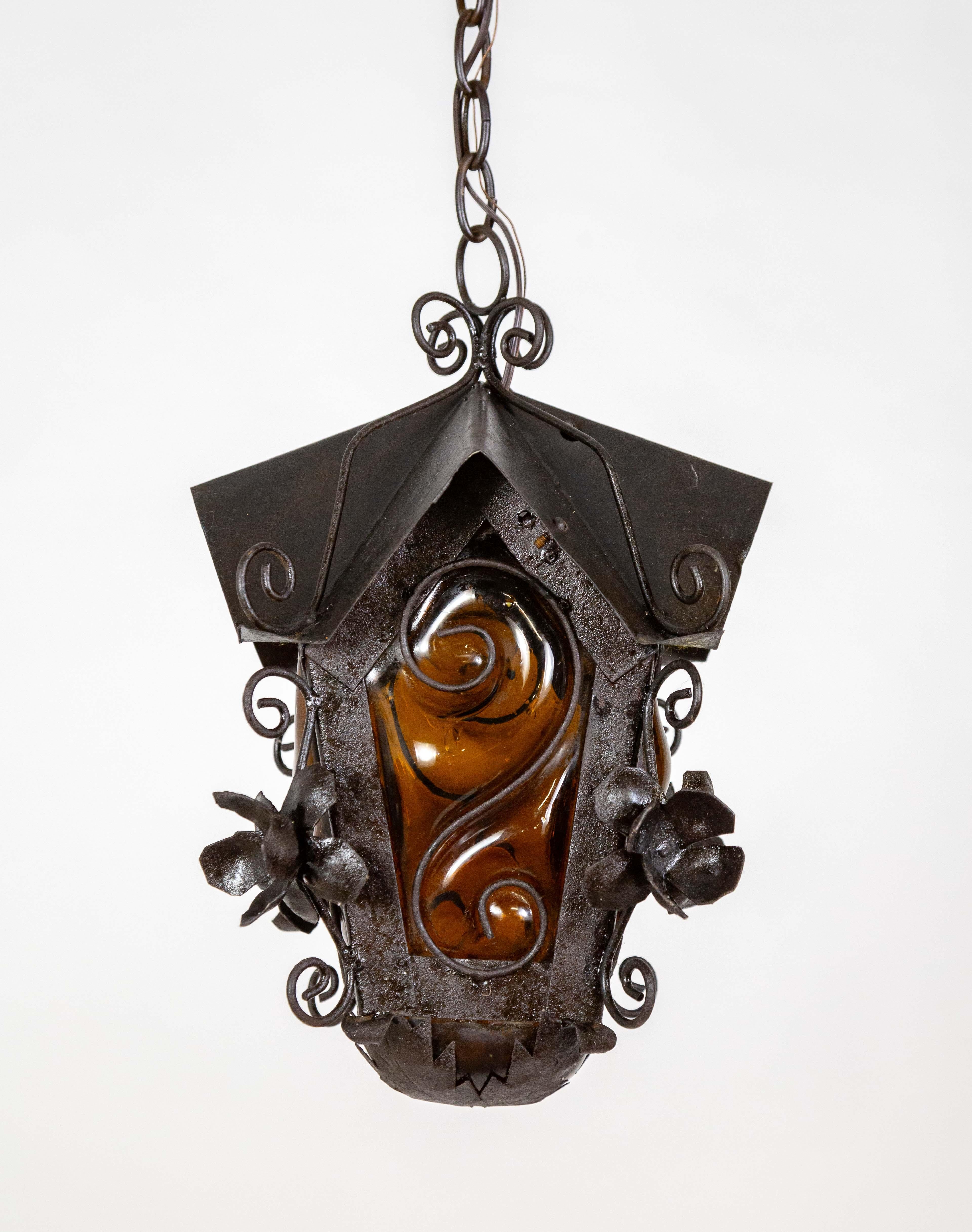 A mid-20th century black metal birdhouse-shaped hanging lantern with a gothic feel.  It is detailed with dimensional flowers and curls between the windows. The four windows are amber blown glass caged in an S-curve. Light also emits from the bottom