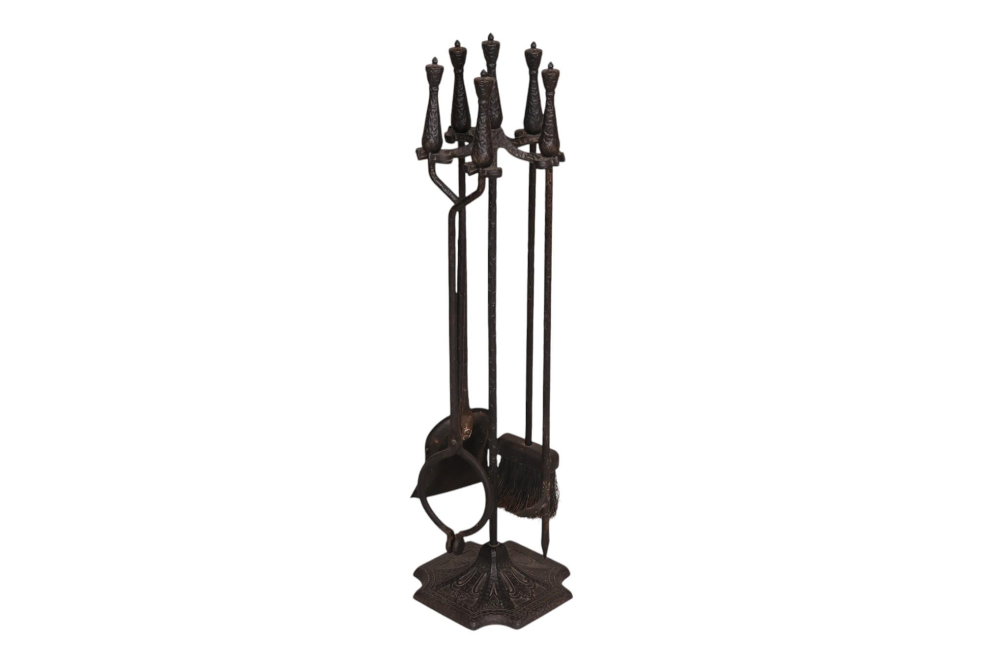 A set of black cast iron fireplace tools. An ash rake, poker, shovel and brush each have a shapely cast iron handle topped with an urn finial. Tools hang from a cast iron yoke with a cast iron base decorated with a simple lily motif. Marked “No 473”