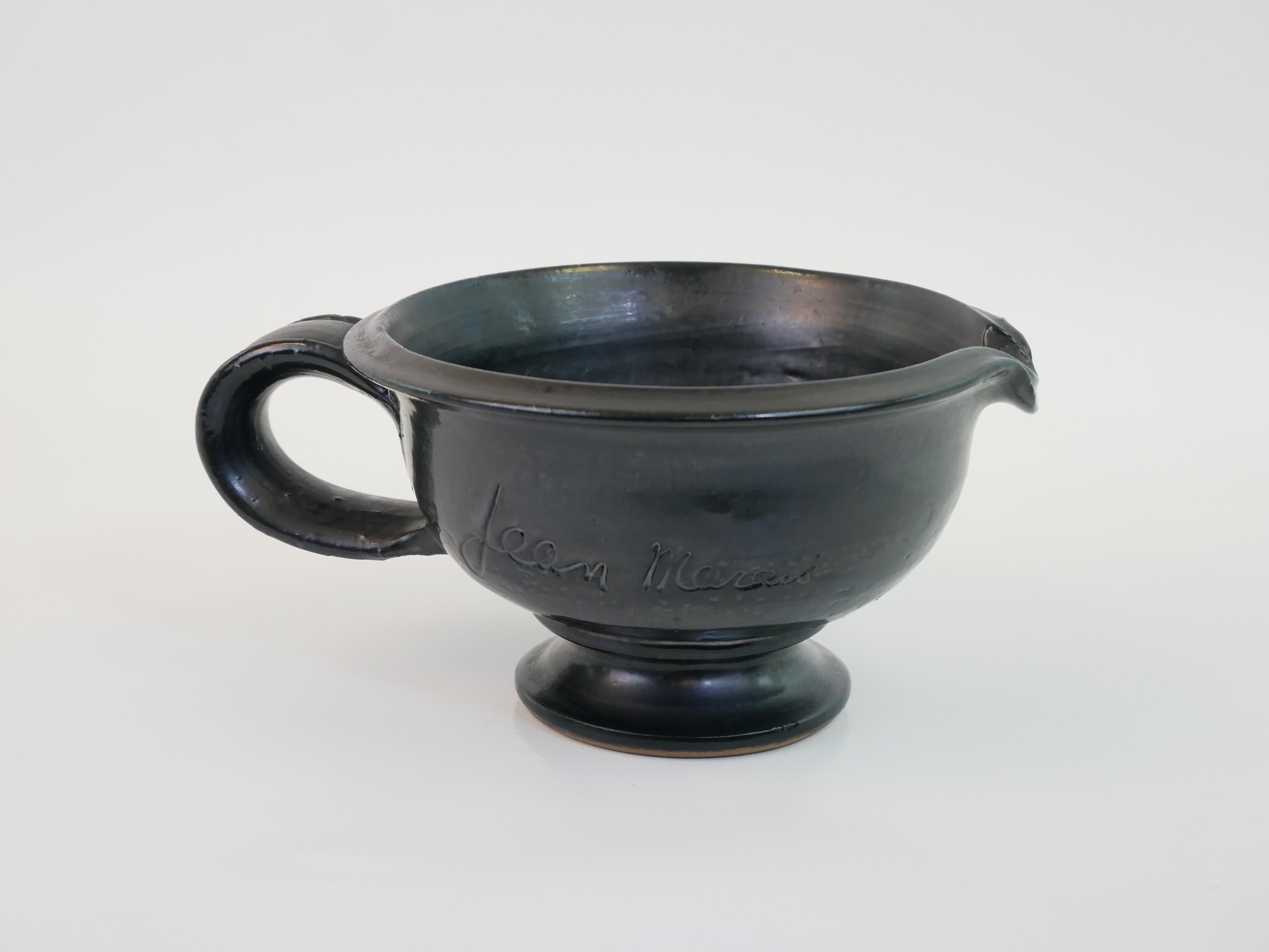 This ceramic pouring vessel, crafted by Jean Marais, showcases a refined black glaze with a smooth, slightly glossy finish. It features a rounded body that elegantly tapers into a pedestal base, ensuring stability. The wide spout is slightly curved,