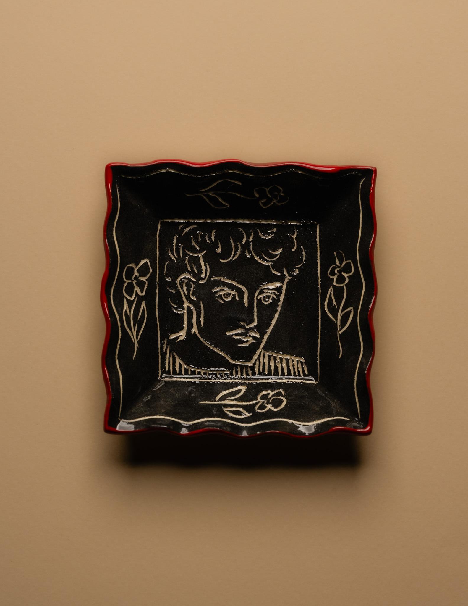 Kleine quadratische Schale aus schwarz glasierter Keramik mit einem eingravierten männlichen Gesicht, umgeben von Blumenmotiven. Einzigartiges Stück, signiert 