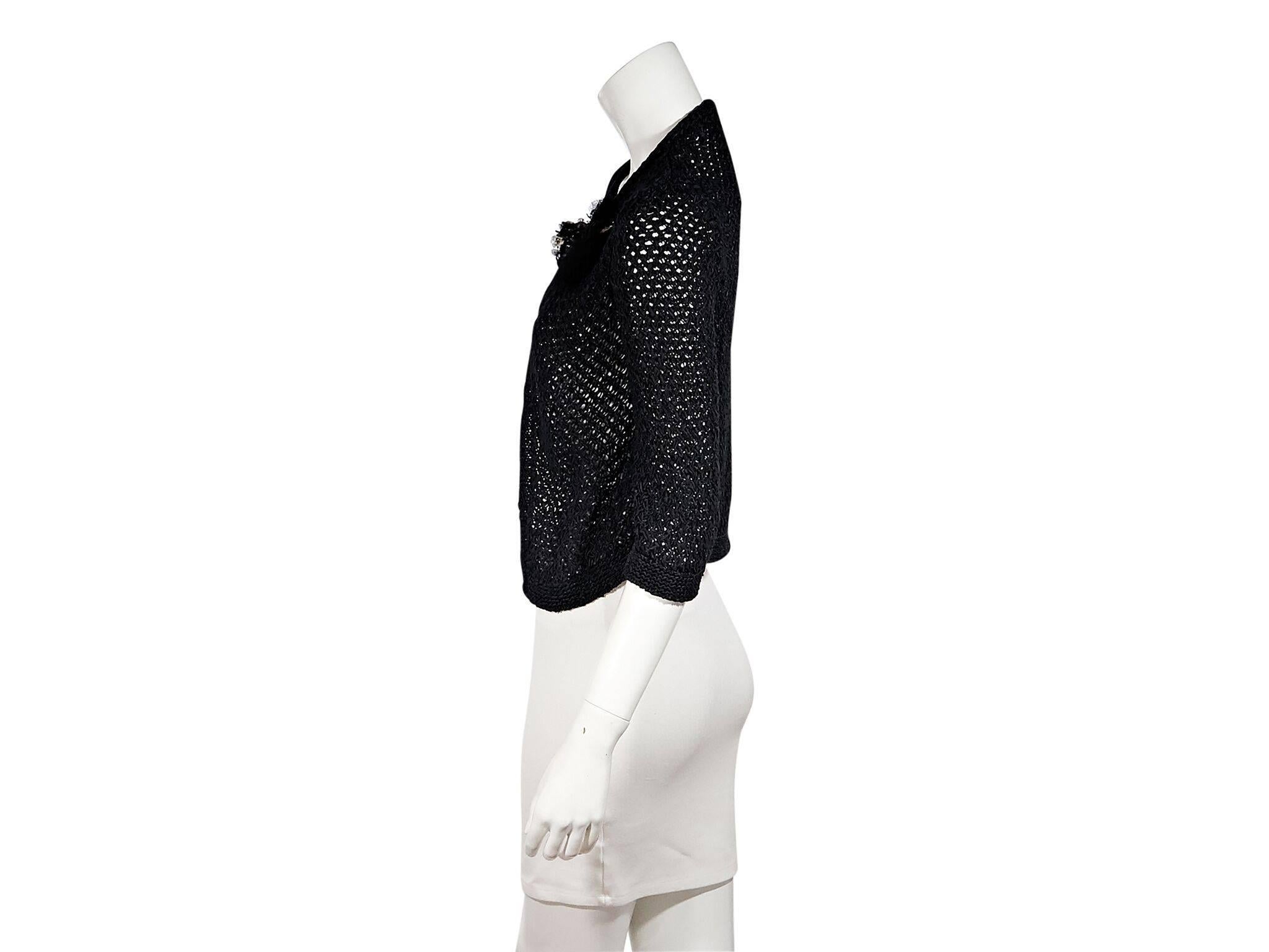 Product details:  Black woven cardigan by Chanel.  Accented with a floral pin.  Elbow-length sleeves.  Chain tie closure.  42